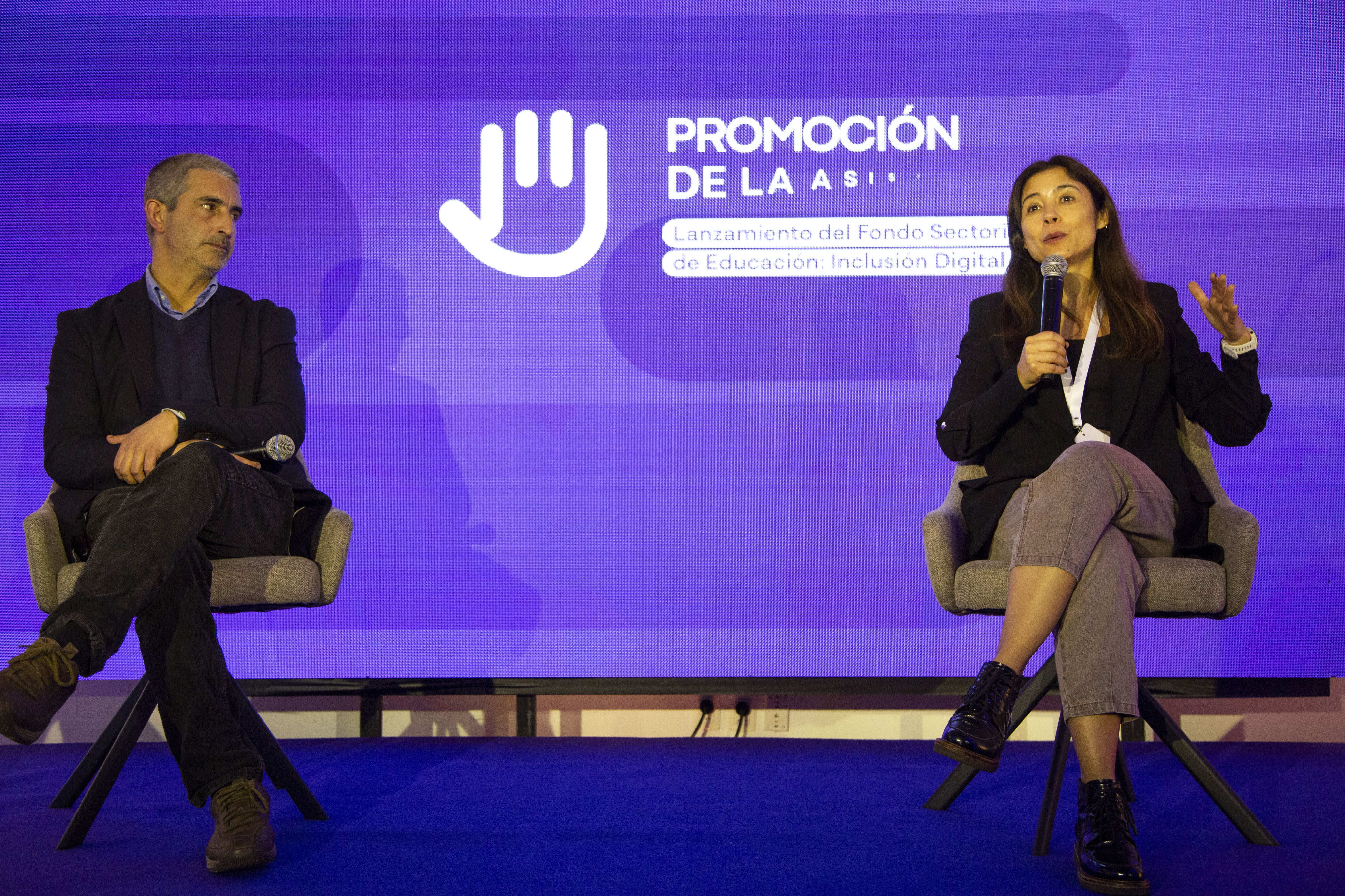 Una mujer diserta sentada en sillón con micrófono en mano, a su costado, el presidente de la ANEP la observa. Atrás, cartel con logo del evento