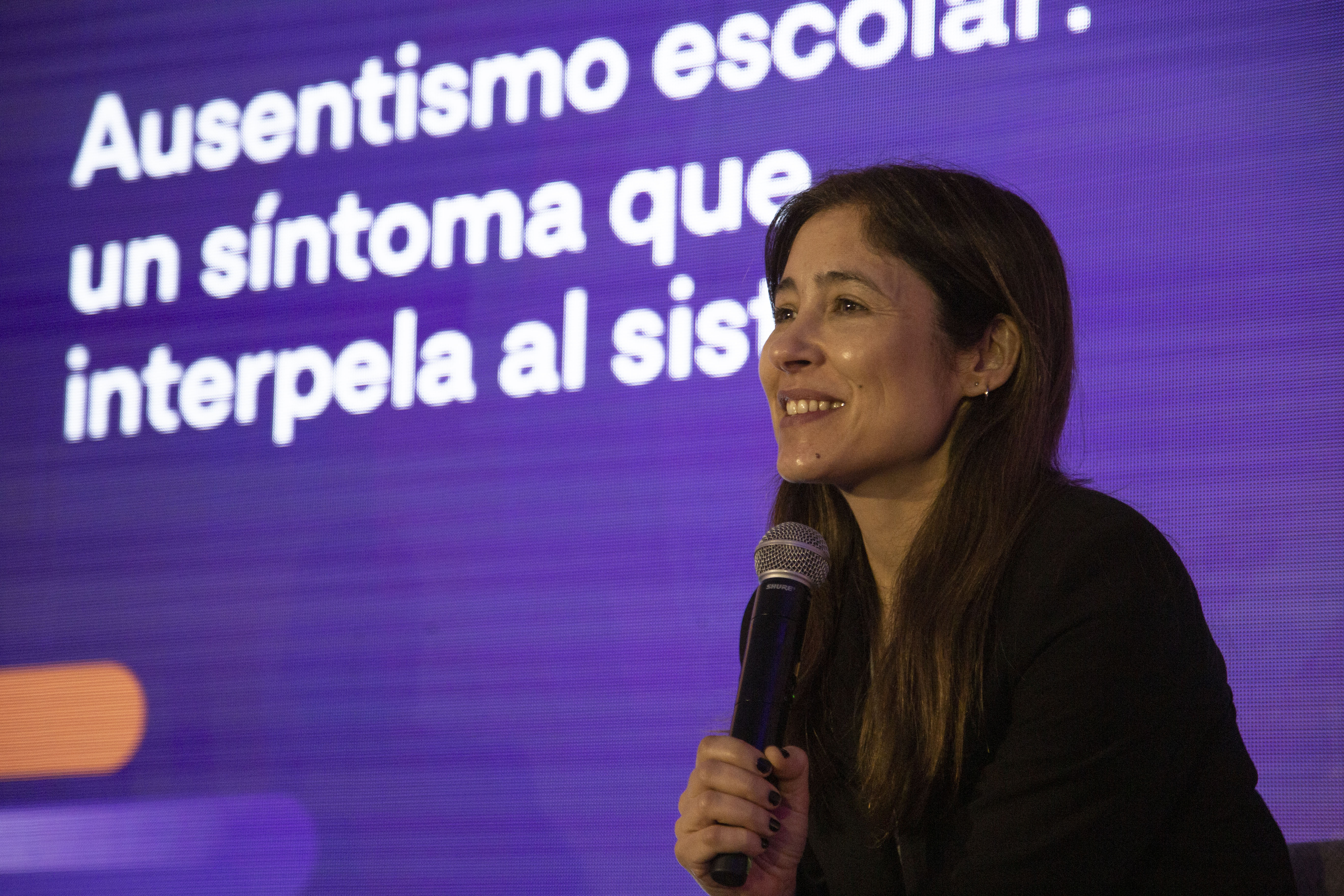 Mujer diserta sentada con micrófono en mano. Atrás, cartel violeta con logo del evento.
