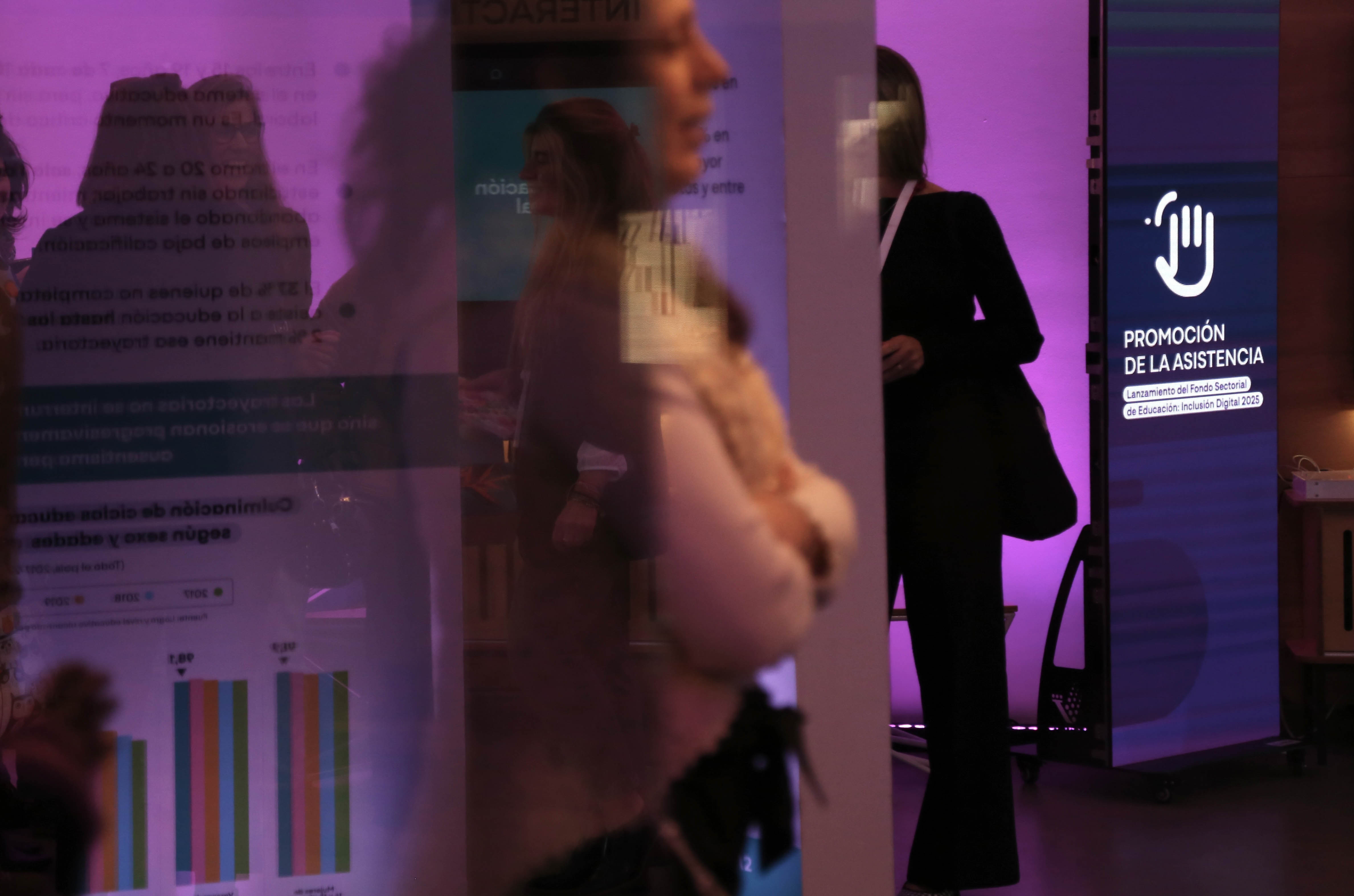 Personas, desenfocadas, paradas en el ingreso a sala de conferencias donde hay carteles con información