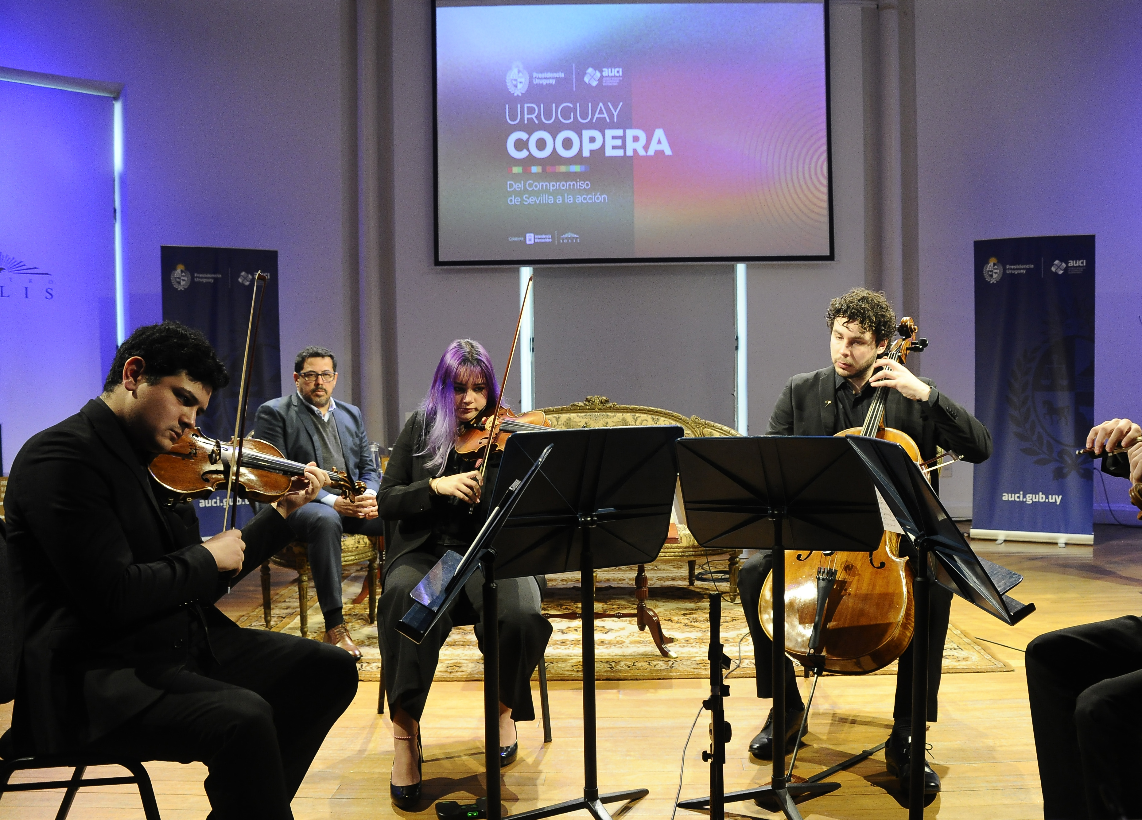 Orquesta Nacional Juvenil del SODRE toca en el escenario. Atrás se observa al secretario de Presidencia sentado observando.