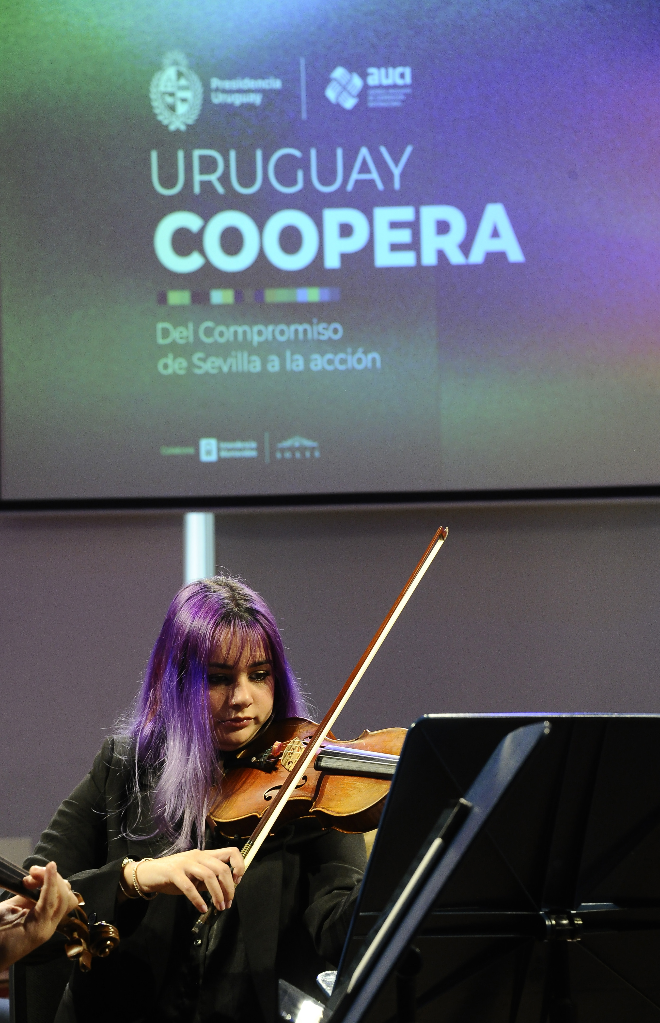 Una mujer toca el violín. Atrás se observa una pantalla con el logo de Uruguay Coopera
