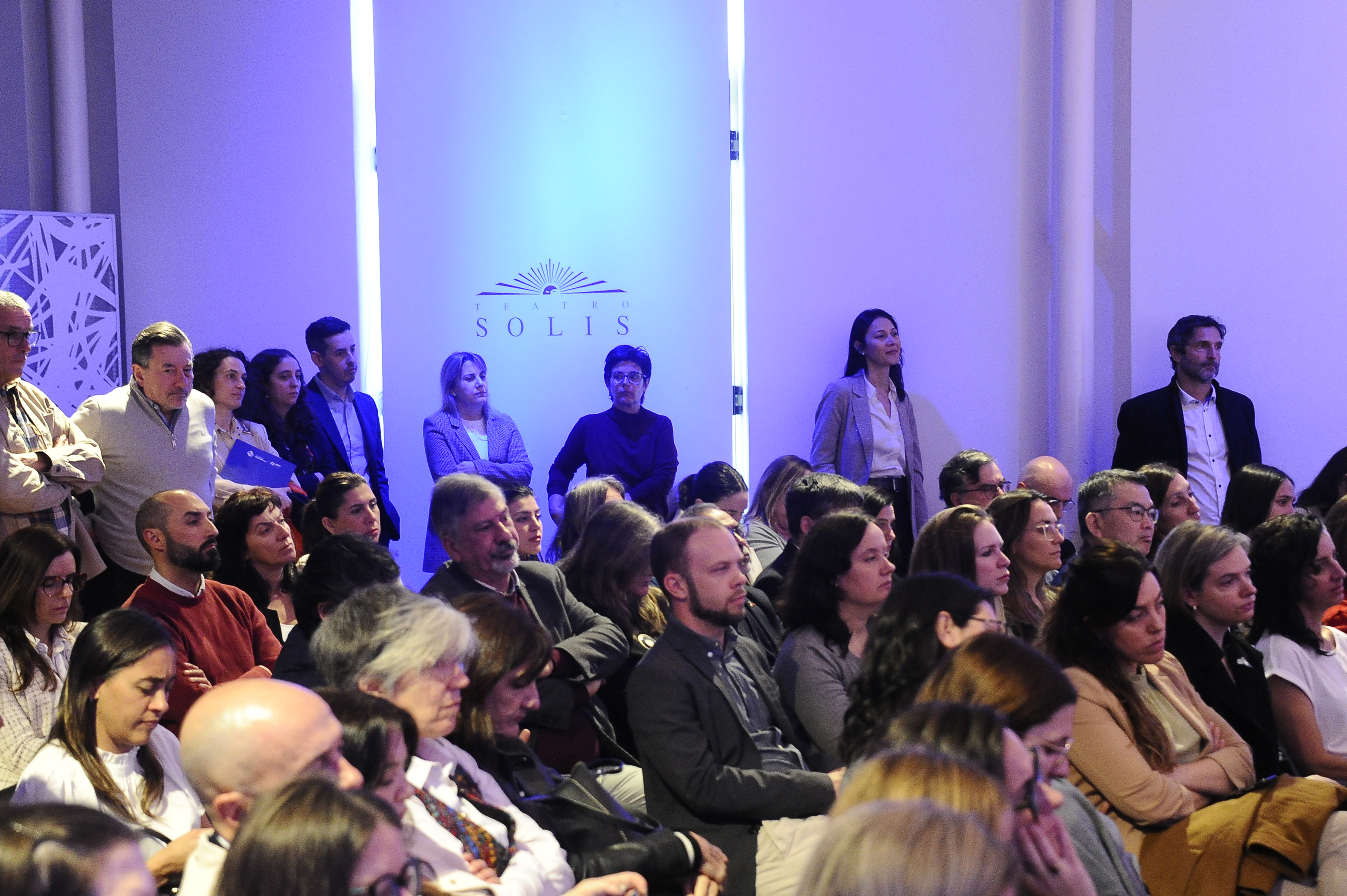 Público sentado en la platea mira hacia el escenario y escucha una disertación