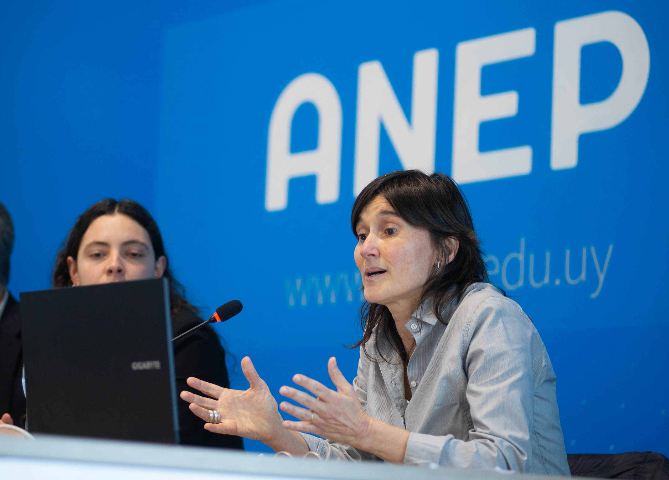 Experta diserta, sentada en mesa con micrófono, en la sala de conferencias de la ANEP. Atrás se observa cartel azul con logo del organismo.