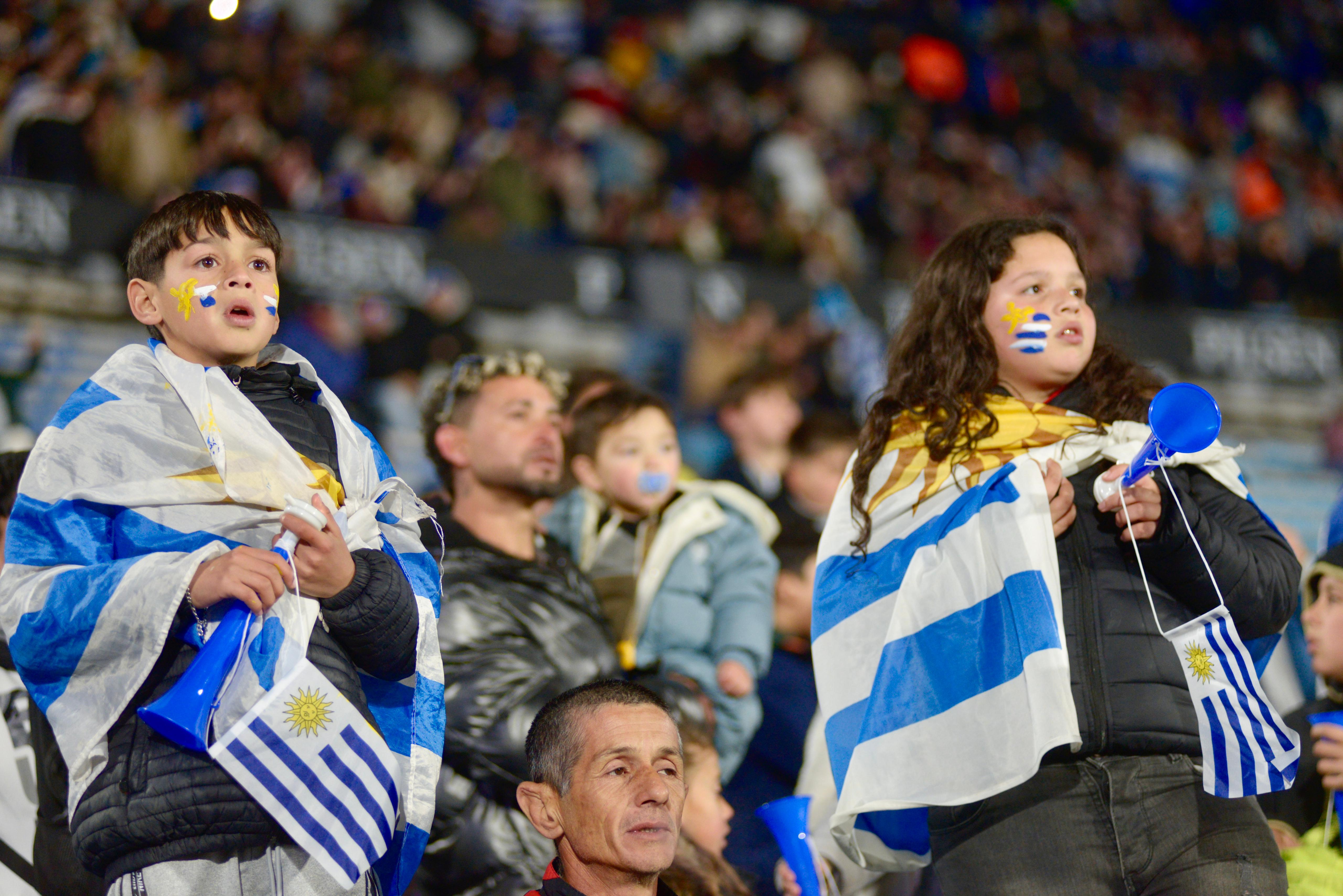Niños y adolescentes parados en la tribuna olímpica del estadio Centenario con globos celestes y blancos y una bandera de Uruguay.