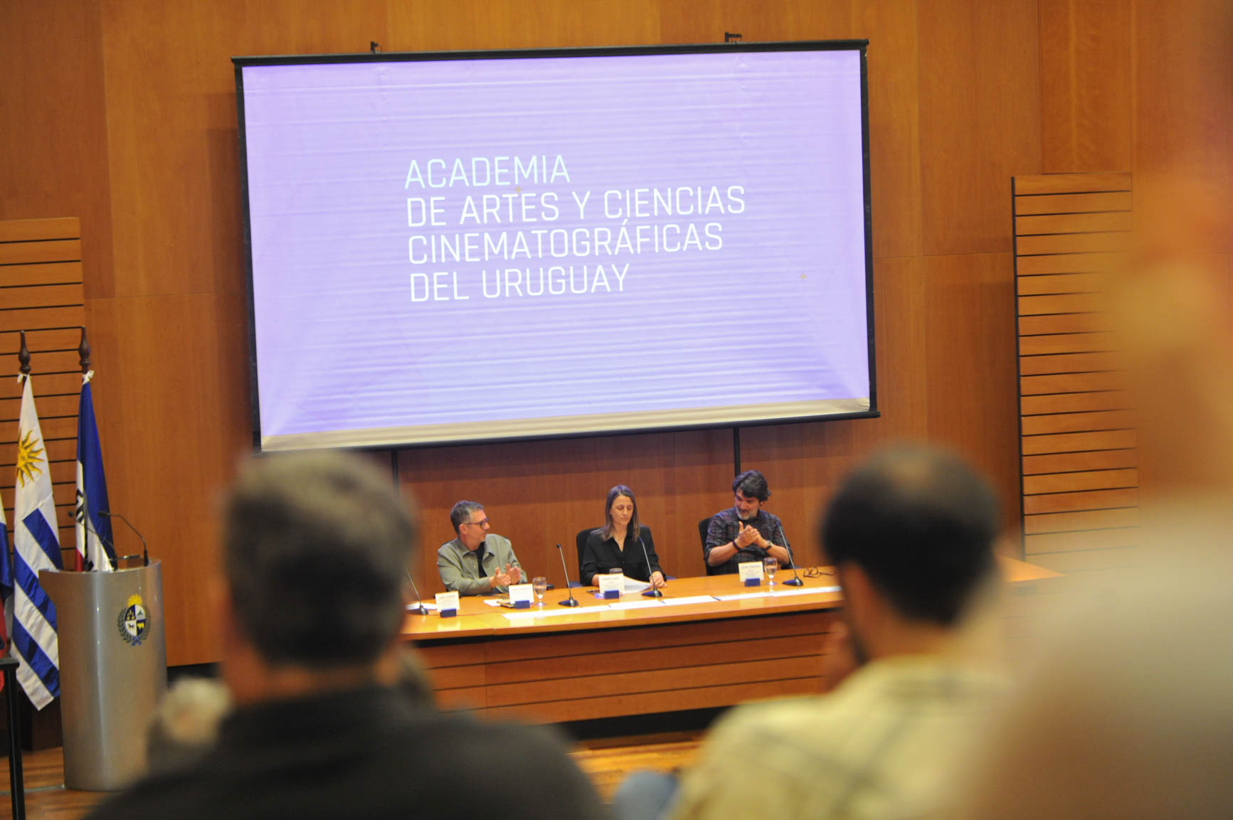 Desde la perspectiva del público, se observa el escenario del auditorio de Presidencia con tres autoridades sentadas disertando.