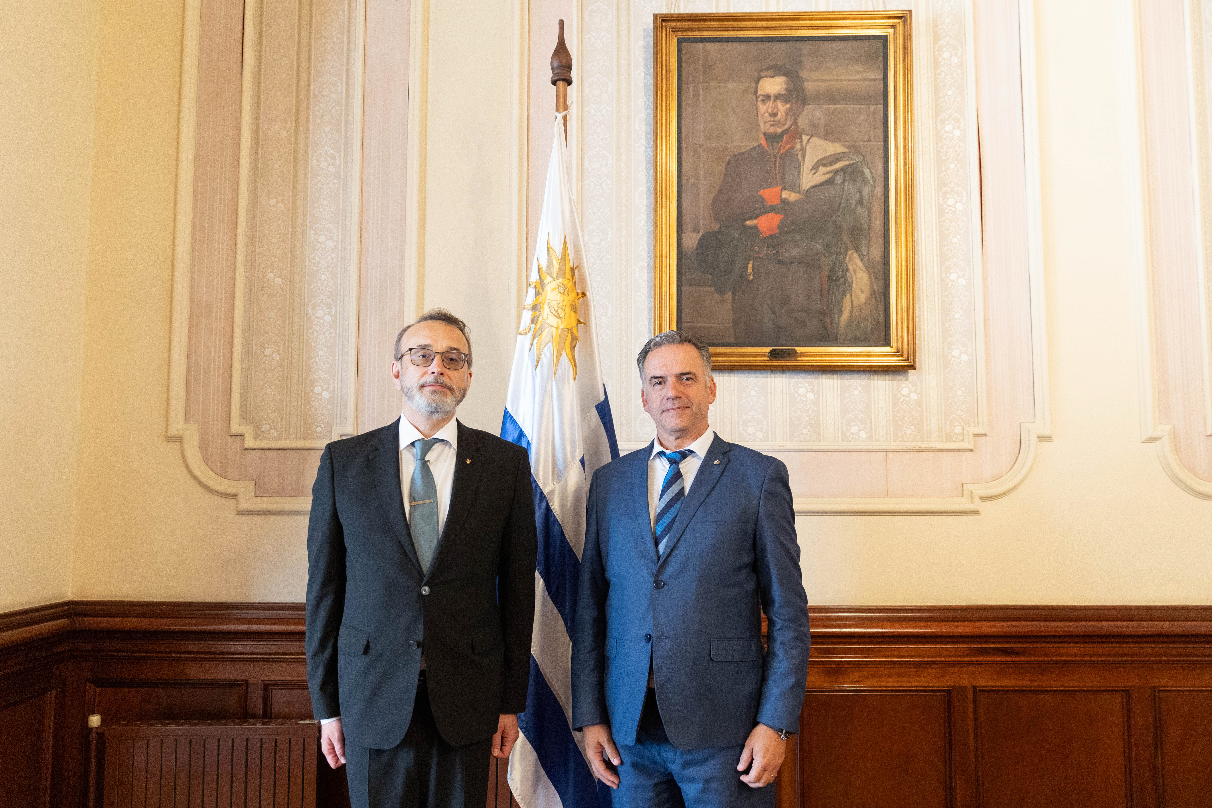 Orsi y el embajador de Ucrania posan sonrientes frente a cámara. Atrás se observa el pabellón nacional y cuadro de Artigas.