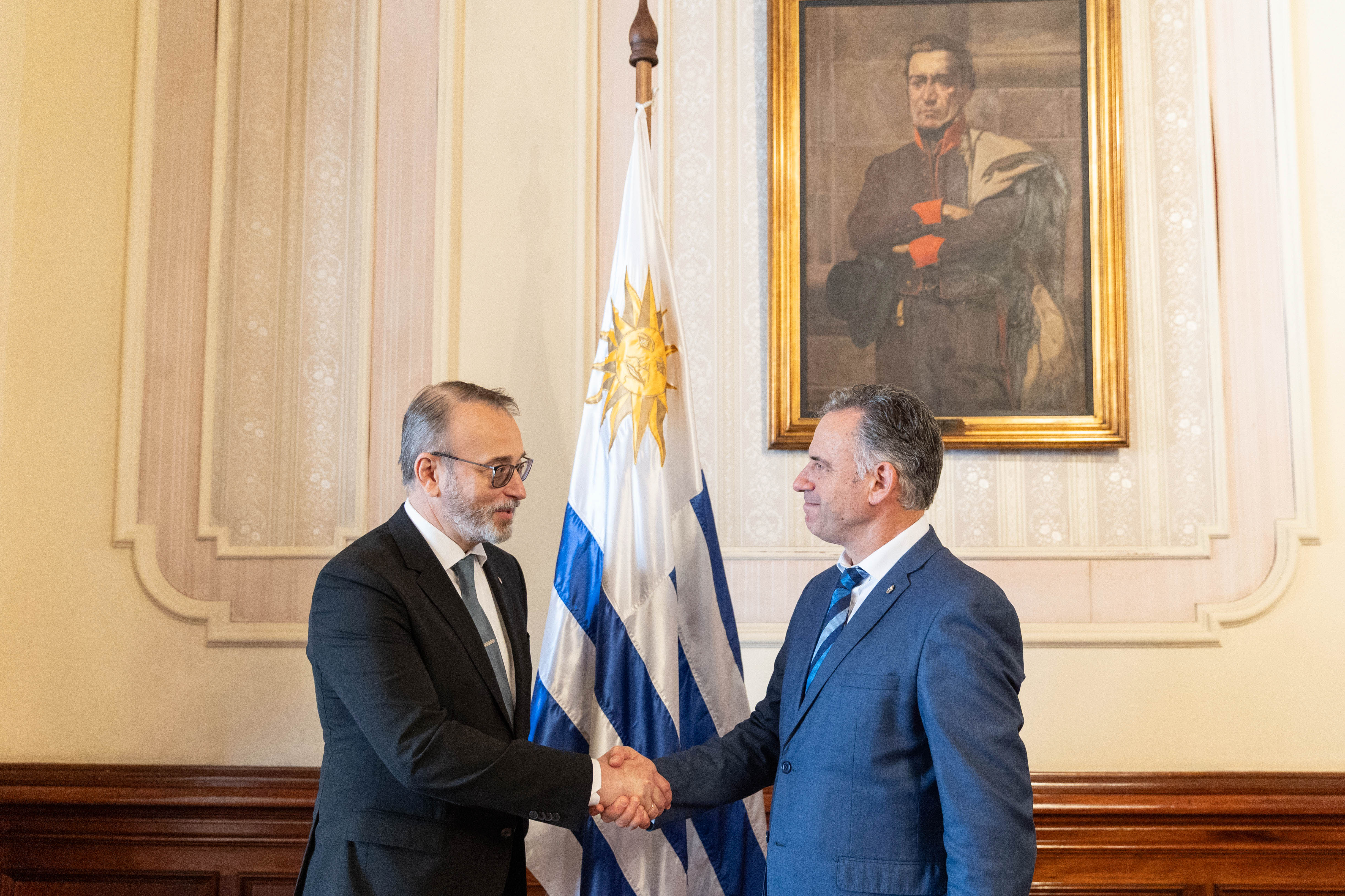 Orsi y el embajador de Ucrania se saludan con la mano. Atrás se observa el pabellón nacional y cuadro de Artigas.