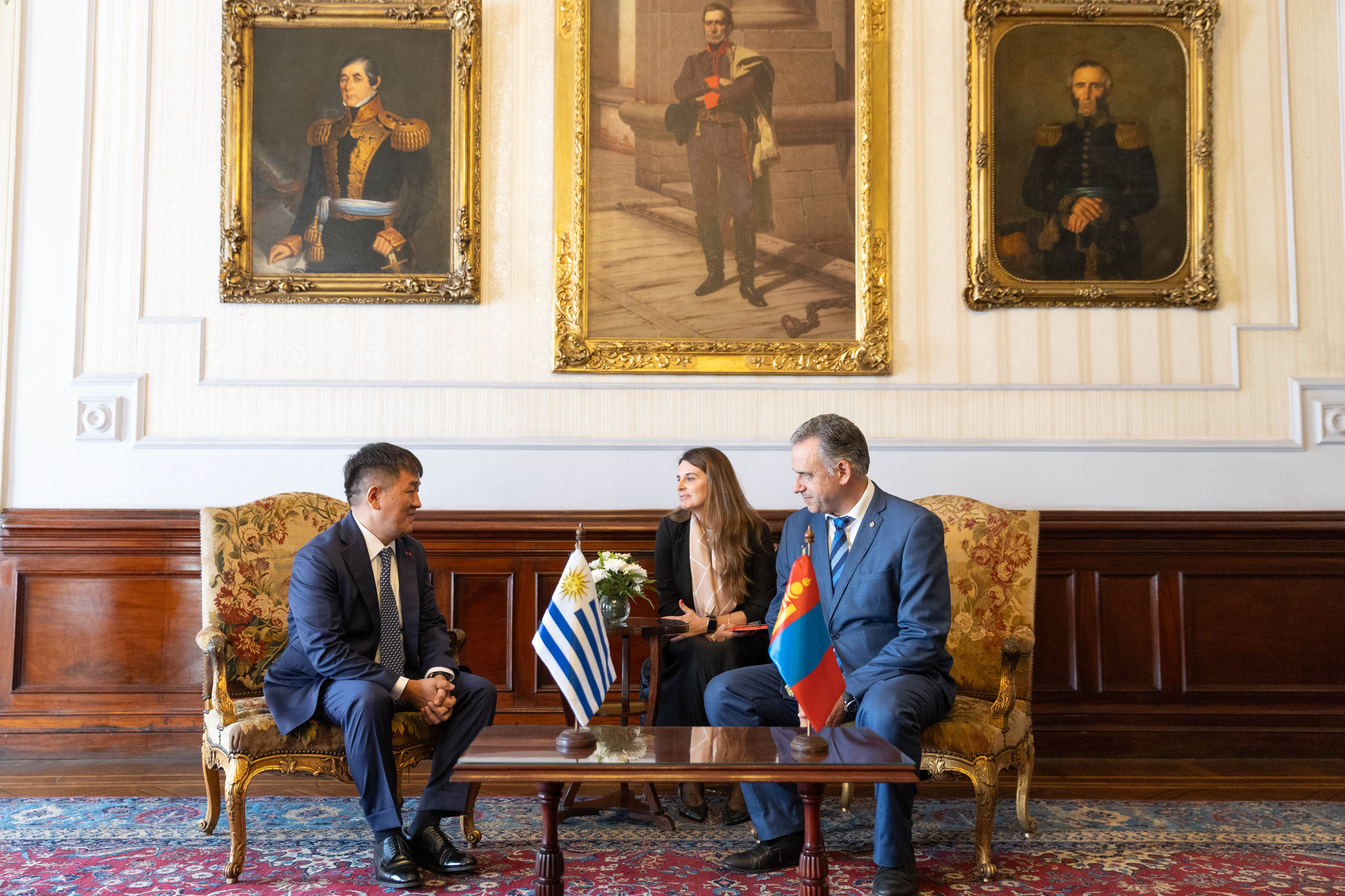 Plano general de Orsi y embajador de Mongolia sentado en sillones dialogando mientras la traductora los observa.