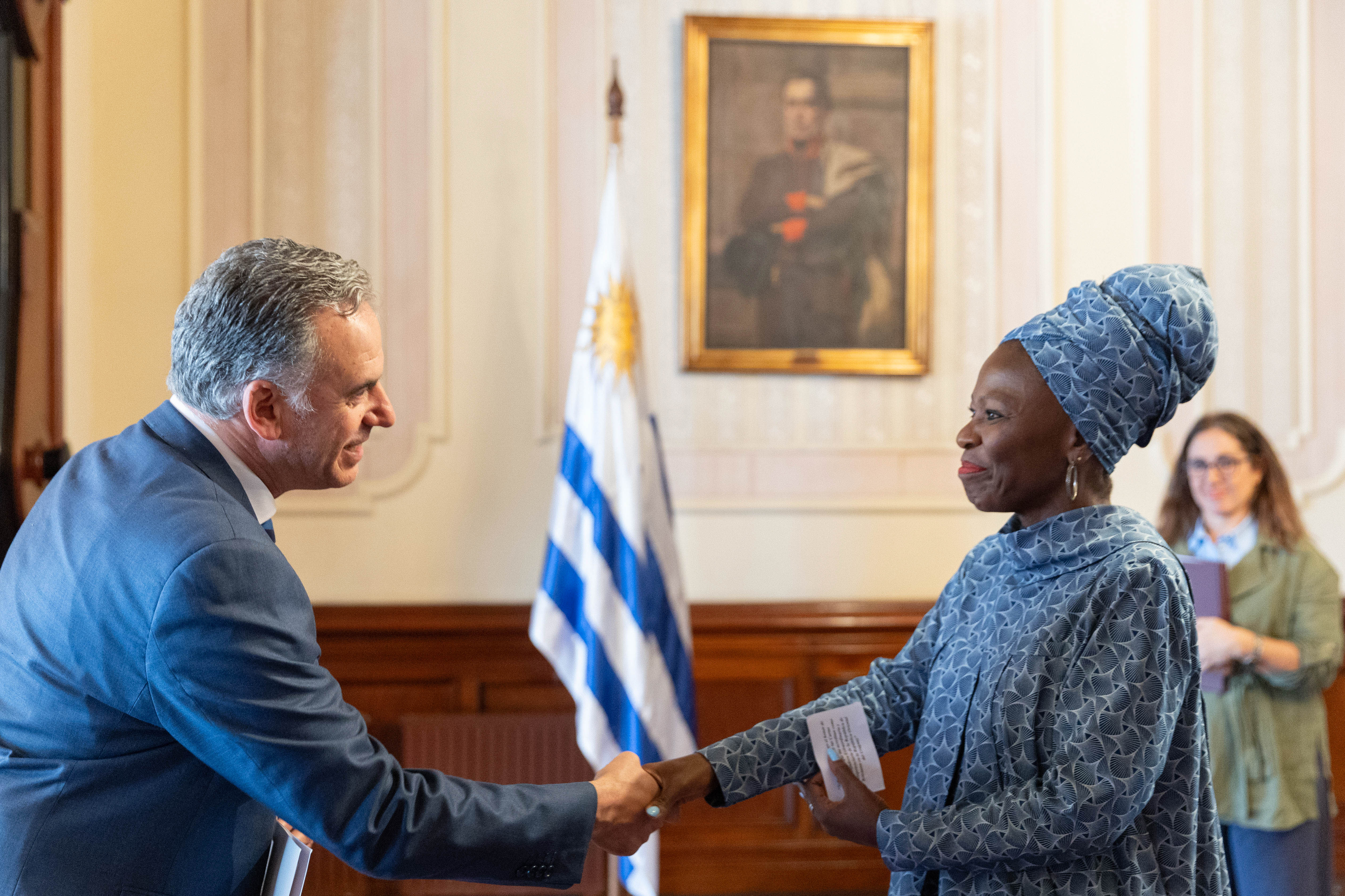 Orsi y embajadora de la República de Botsuana se saludan con la mano en el salón principal. Atrás se observa el pabellón nacional y cuadro de Artigas.