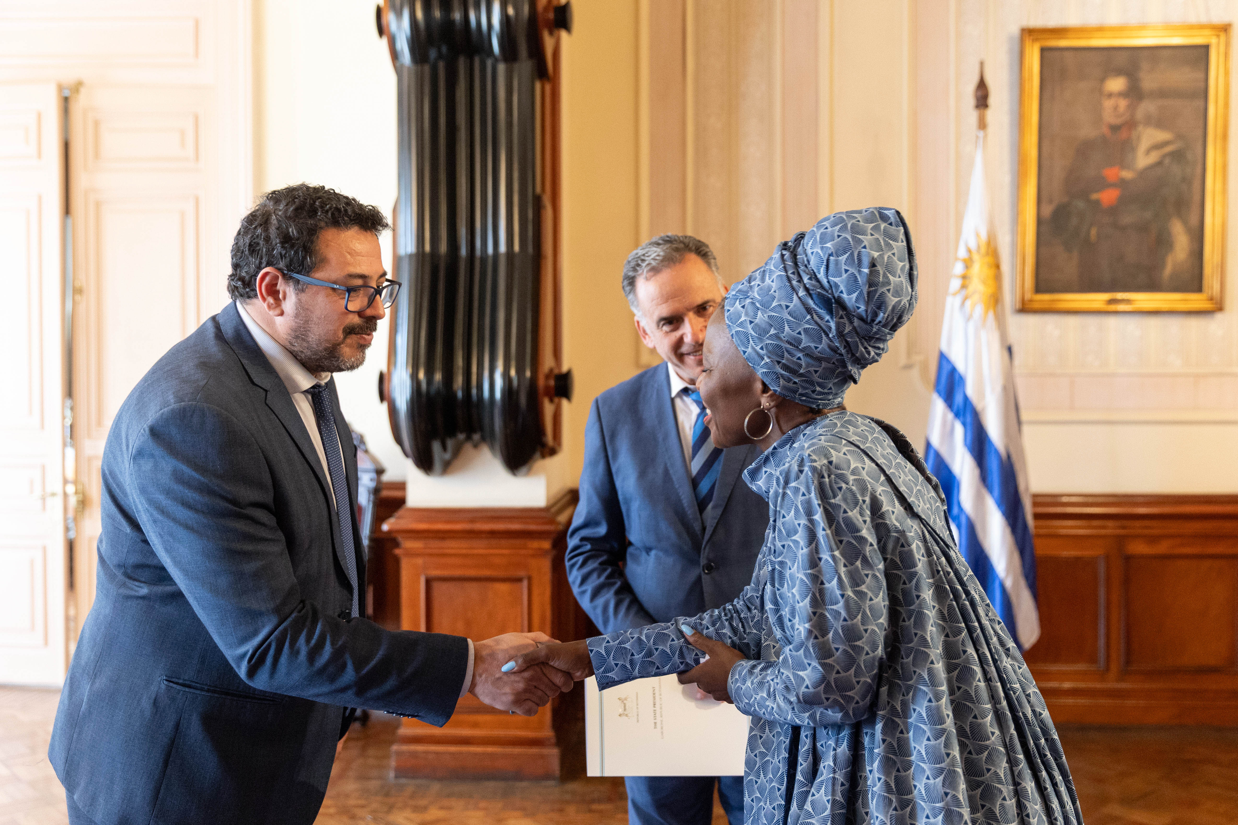 Secretario de Presidencia y embajadora de la República de Botsuana se saludan con la mano mientras Orsi los observa.