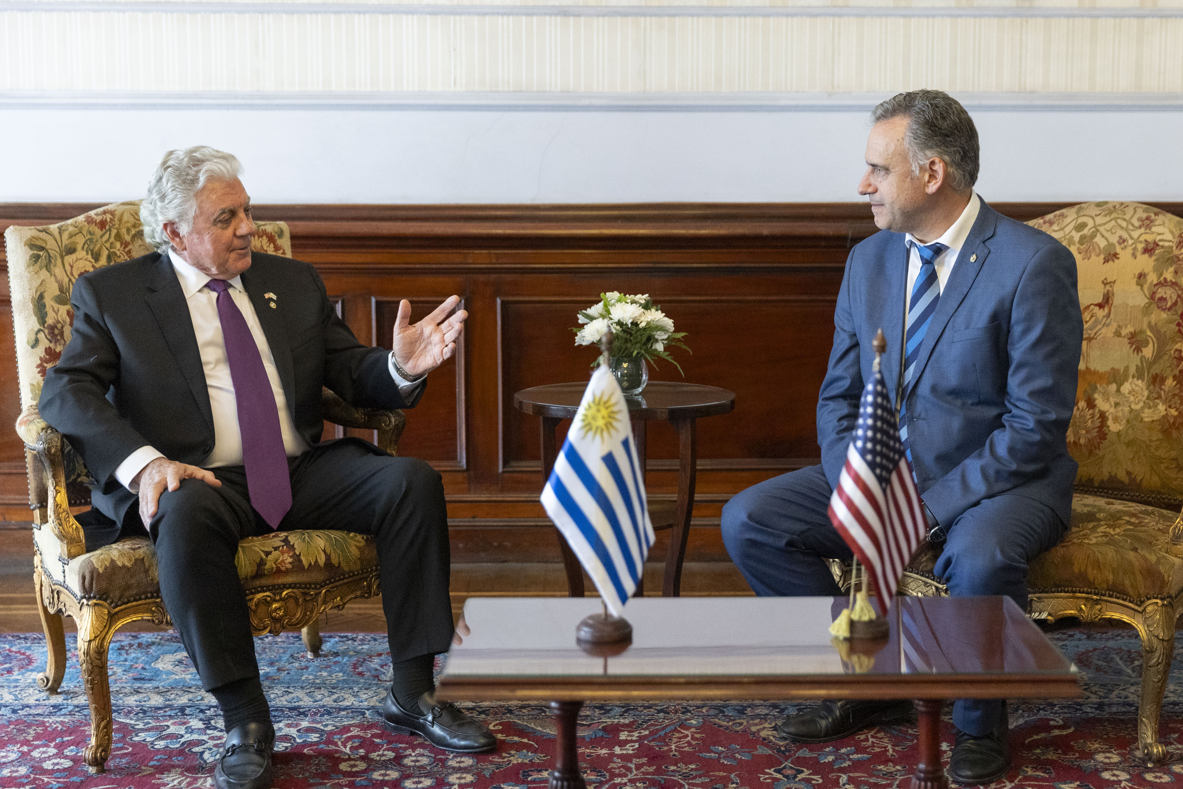 Orsi y embajador de Estados Unidos sentados en sillones dialogan entre sí. Adelante hay una mesa pequeña con banderas de ambos países.