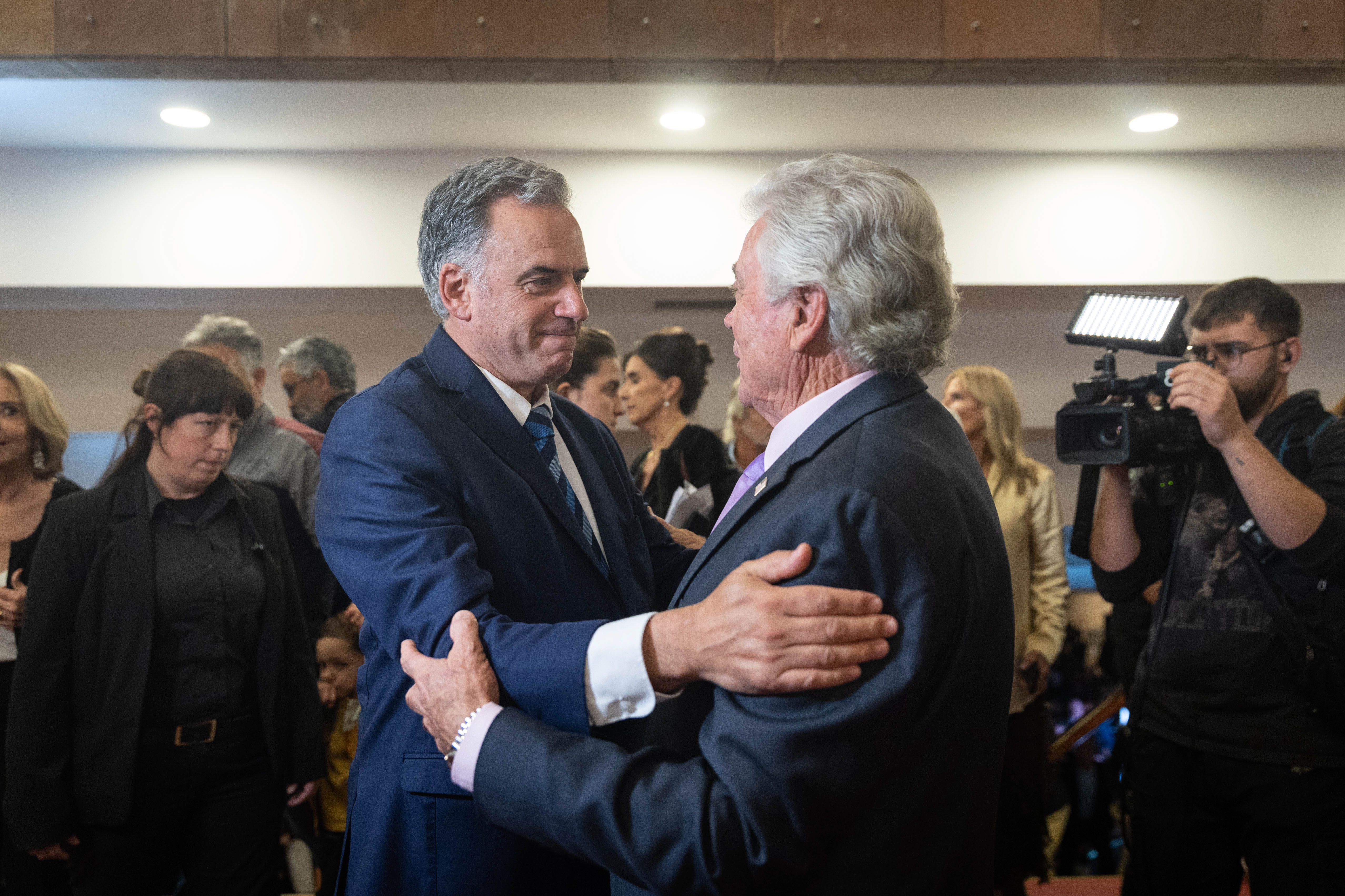 Orsi saluda con un abrazo a un hombre en el ingreso al auditorio. Alrededor camarógrafos y otros asistentes los observan.