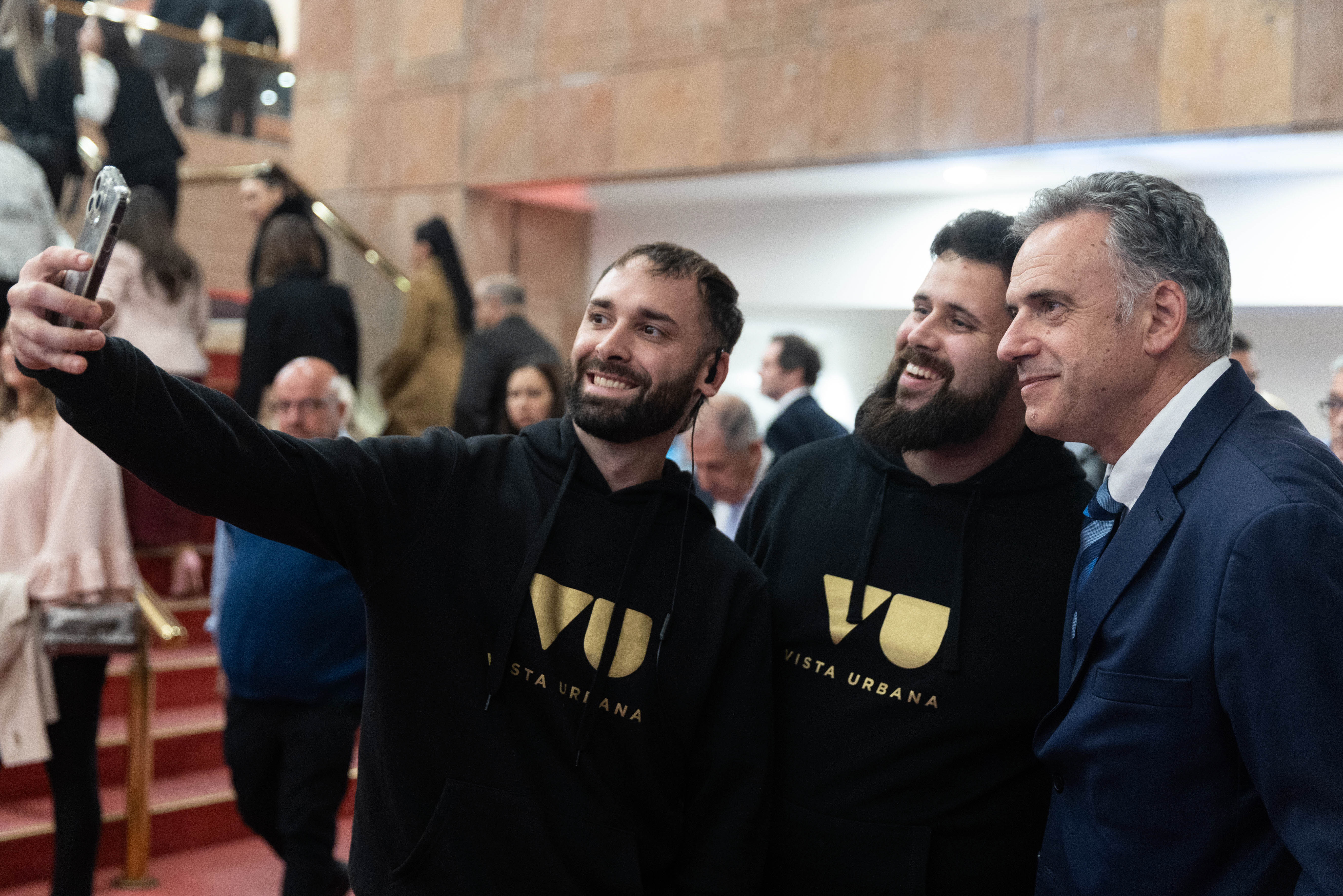 Orsi junto a dos hombres con buzos negros y letras doradas se toman una foto con un celular en el ingreso al auditorio.