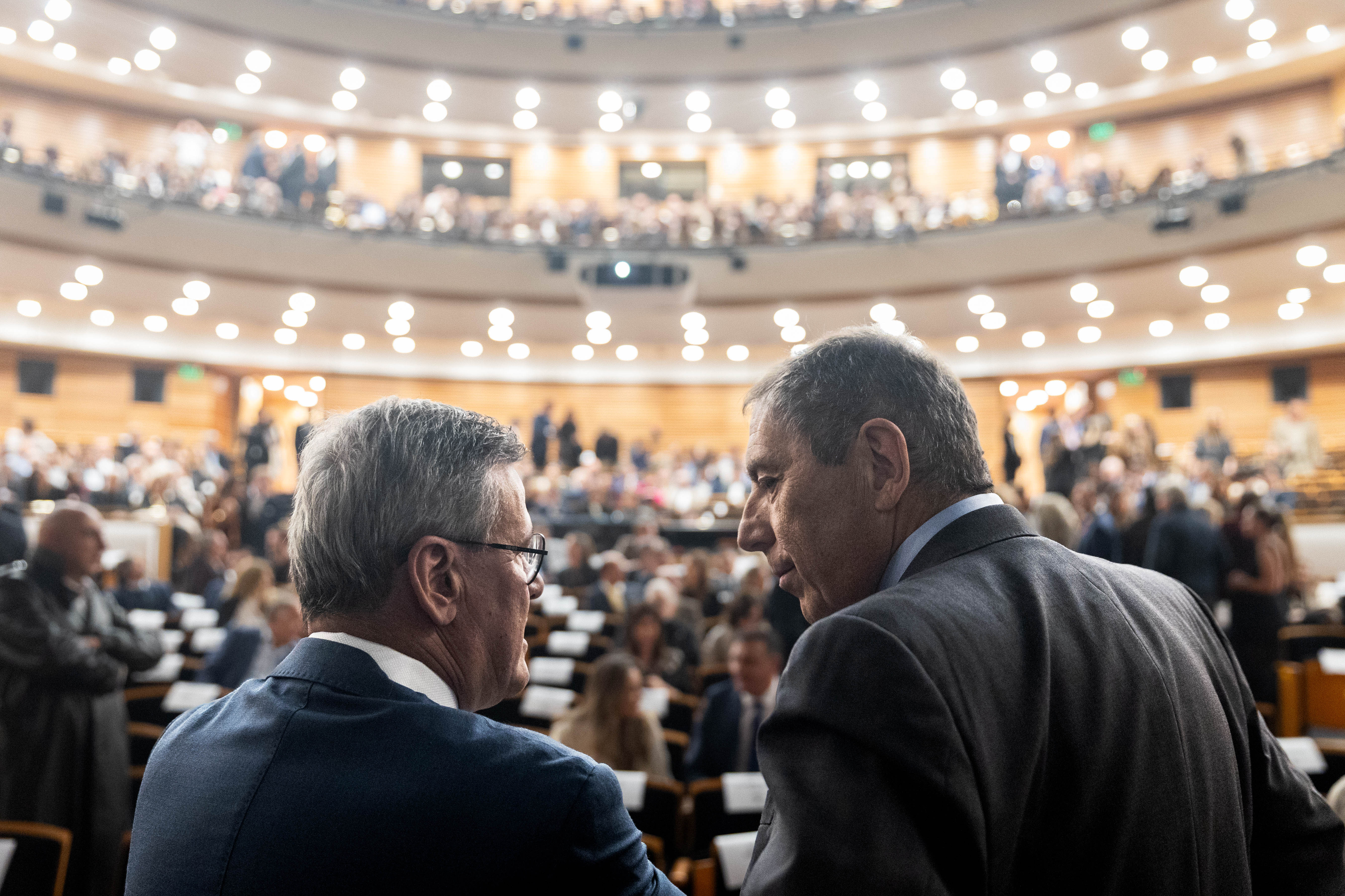 Prosecretario de Presidencia y ministro de Economía dialogan, de espaldas, en la sala Adela Reta. Atrás se observa el público sentado. 