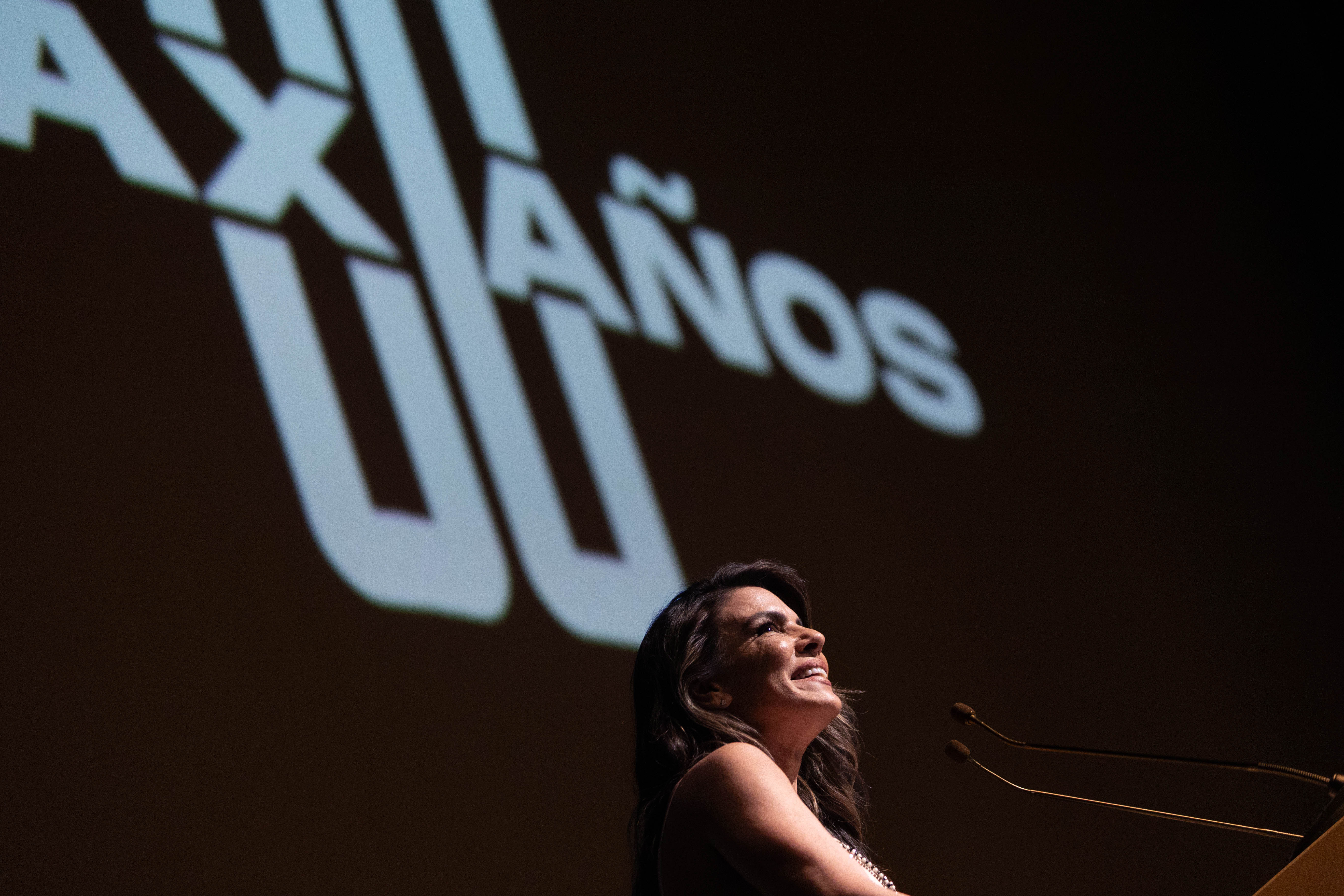 Maestra de ceremonia, sonriente, diserta parada en atril en el escenario. Atrás se observa borroso una proyección con el texto 80 años.