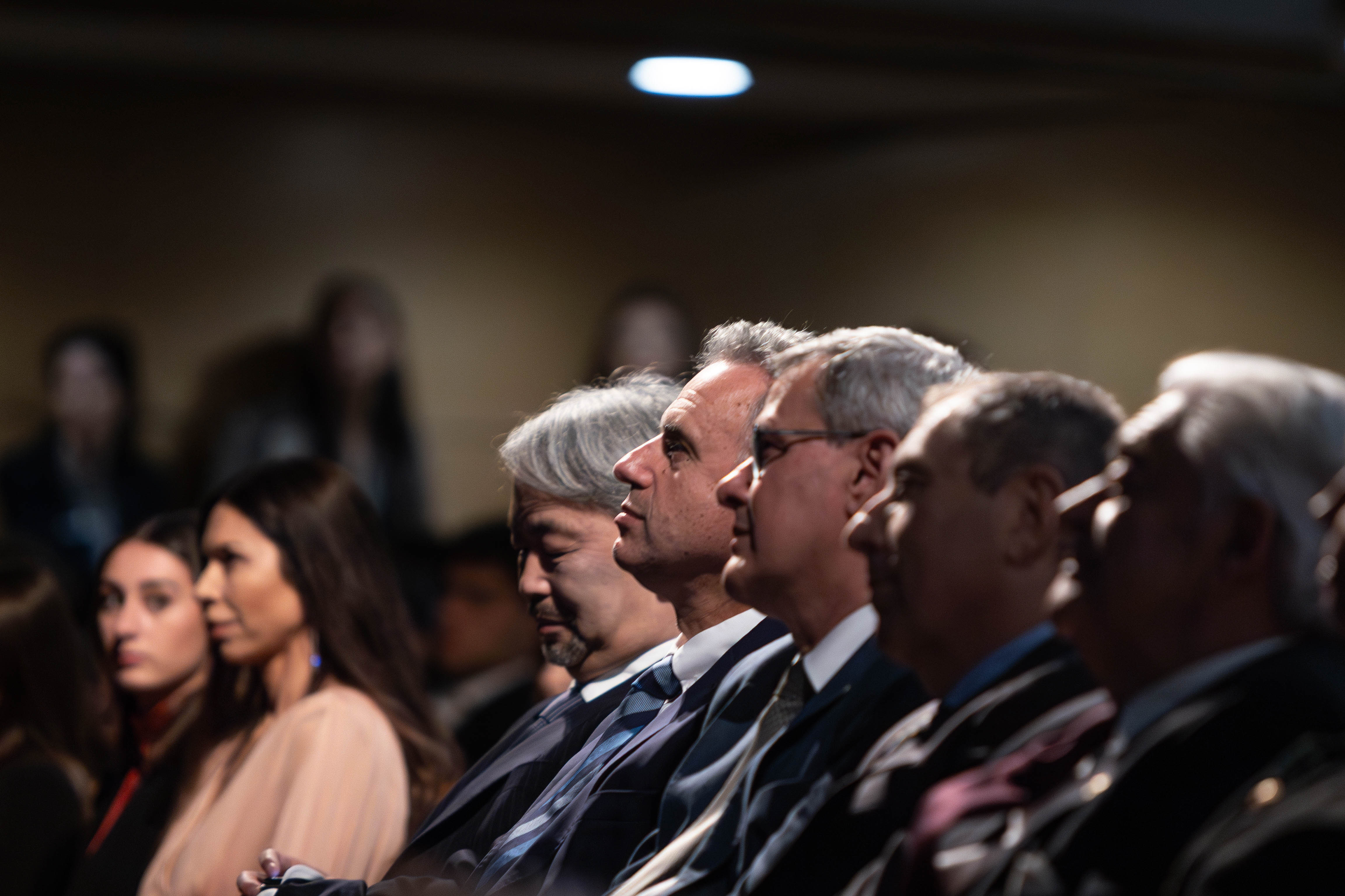 Orsi junto a autoridades nacionales y otros asistentes sentado en primera fila de la platea del auditorio mira hacia el escenario.
