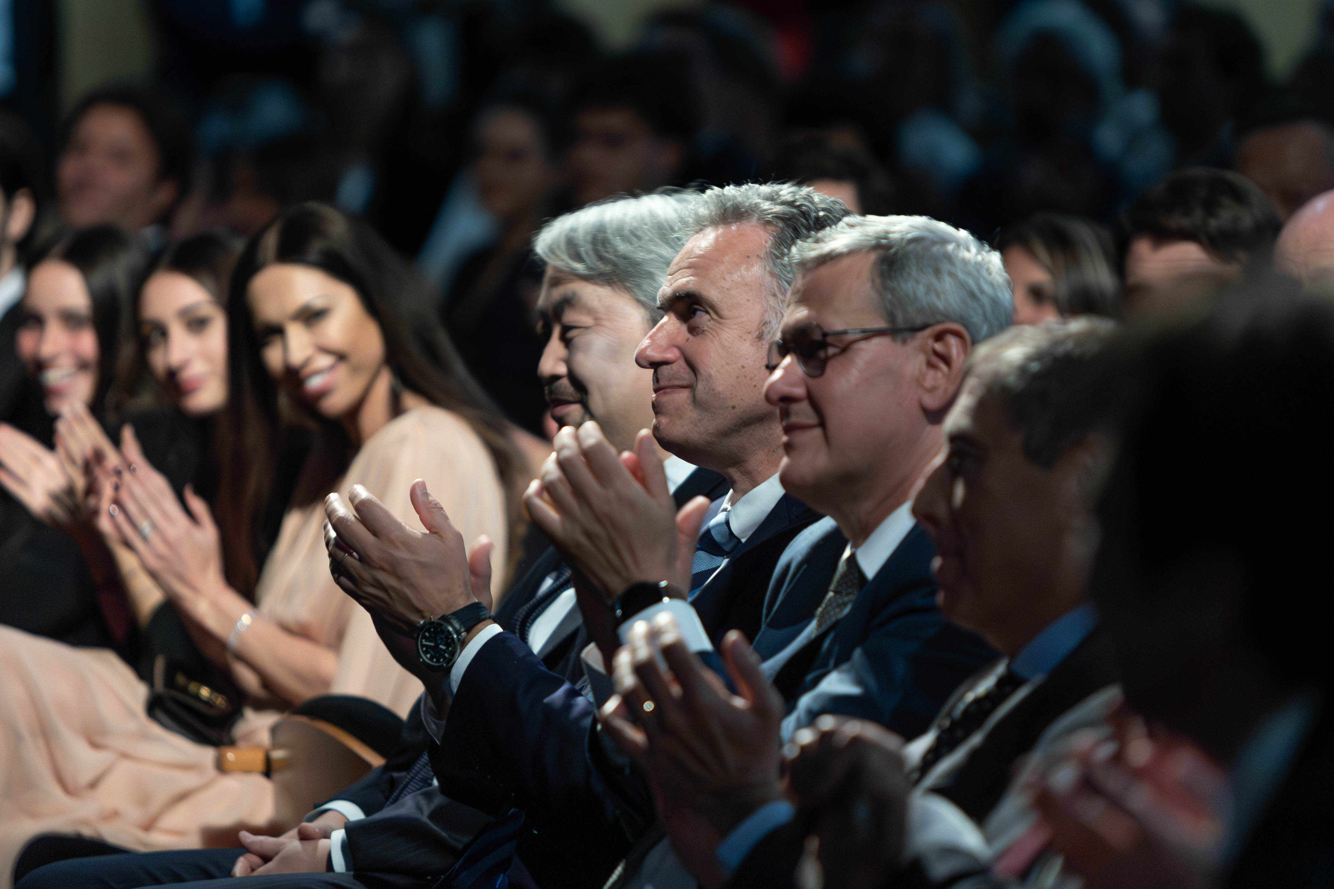 Orsi junto a autoridades nacionales y otros asistentes sentado en primera fila de la platea aplaude mientras mira hacia el escenario.
