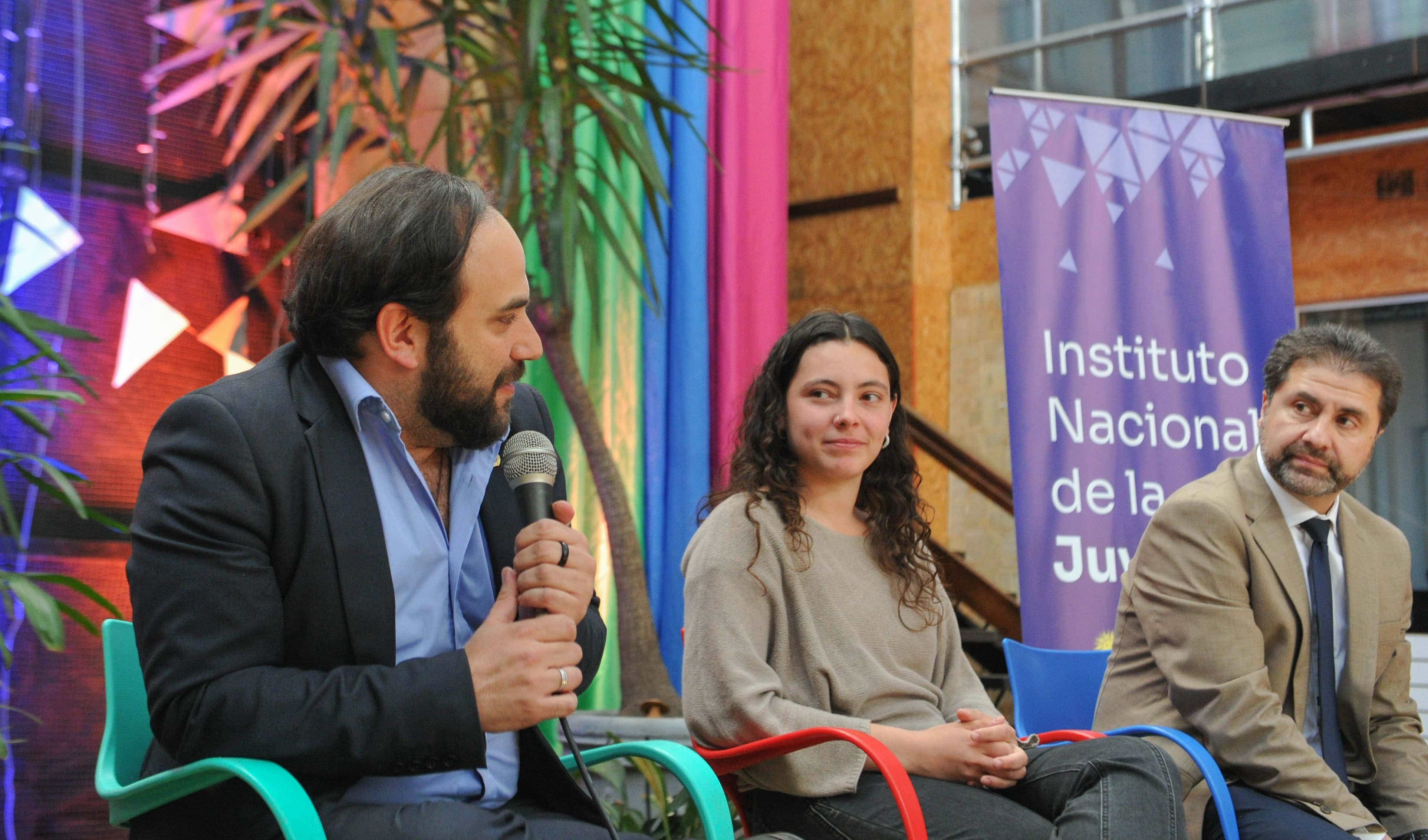 Ministro de Desarrollo Social diserta sentado en silla con micrófono en mano. A su costado está la directora del INJU y representante de atletas.
