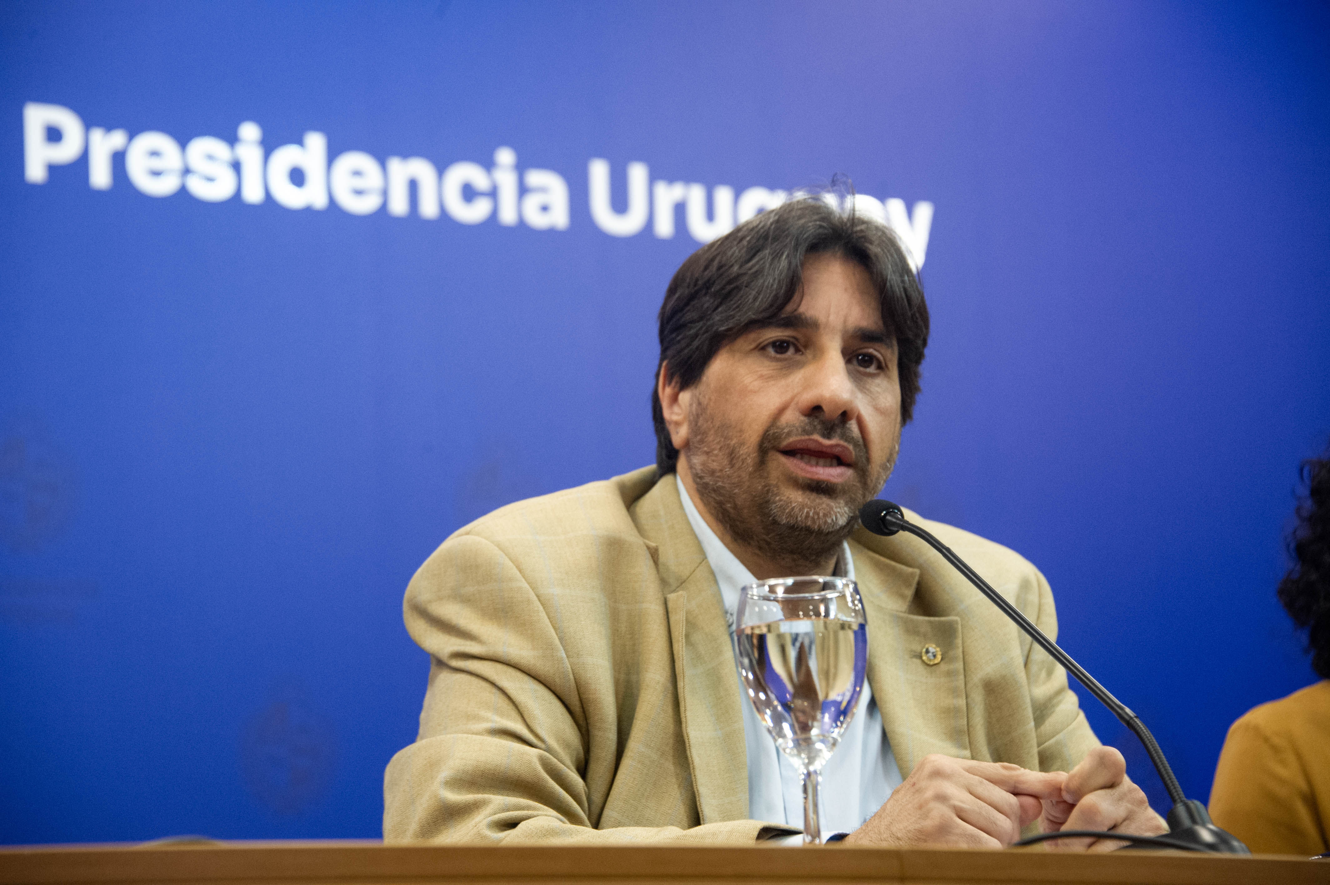 Presidente de la Unasev diserta sentado en mesa con micrófono, en sala de conferencia de Presidencia. Atrás se observa cartel azul con logo.