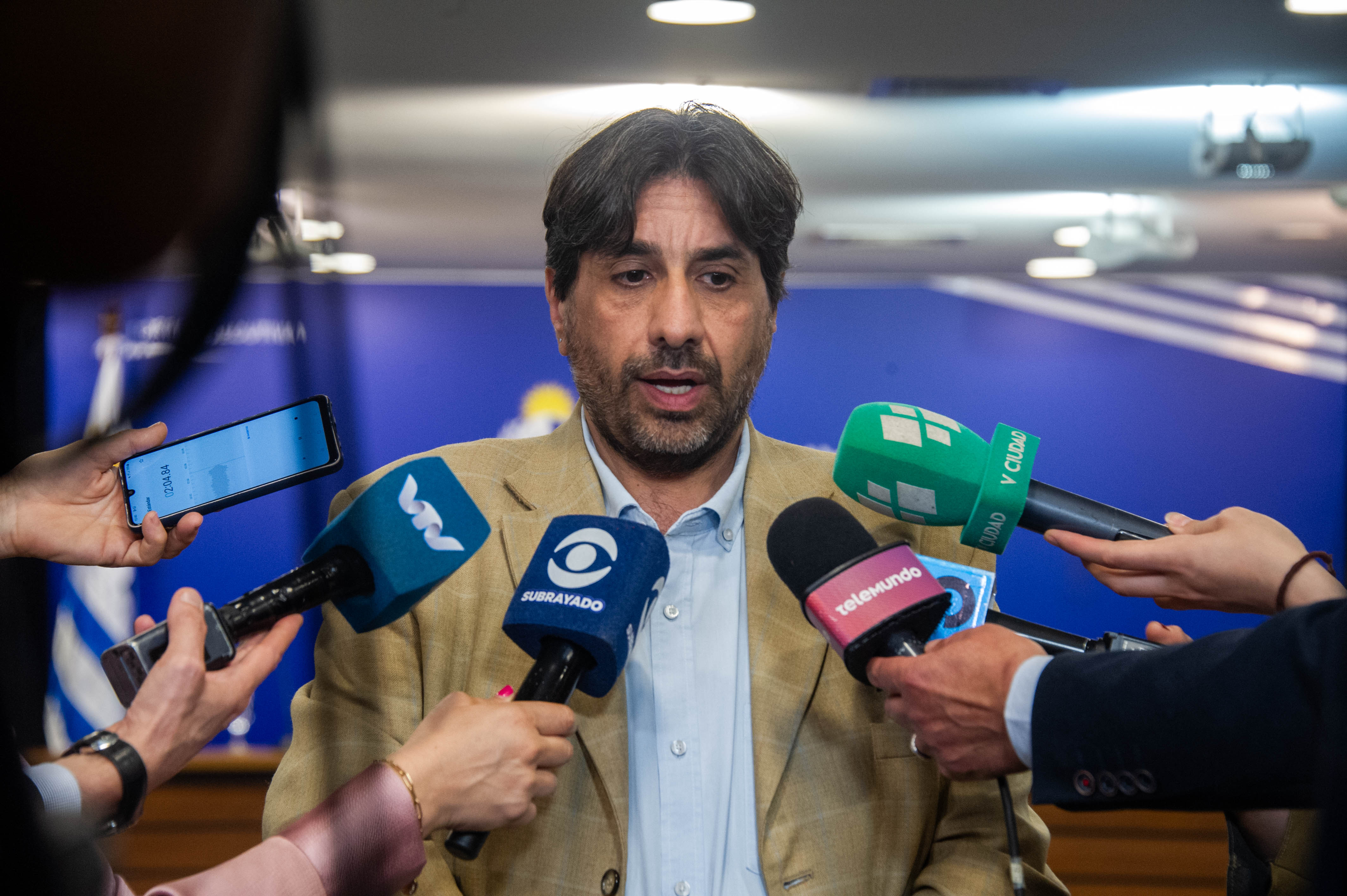 Presidente de la Unasev, de pie, dialoga con periodistas frente a varios micrófonos de prensa.