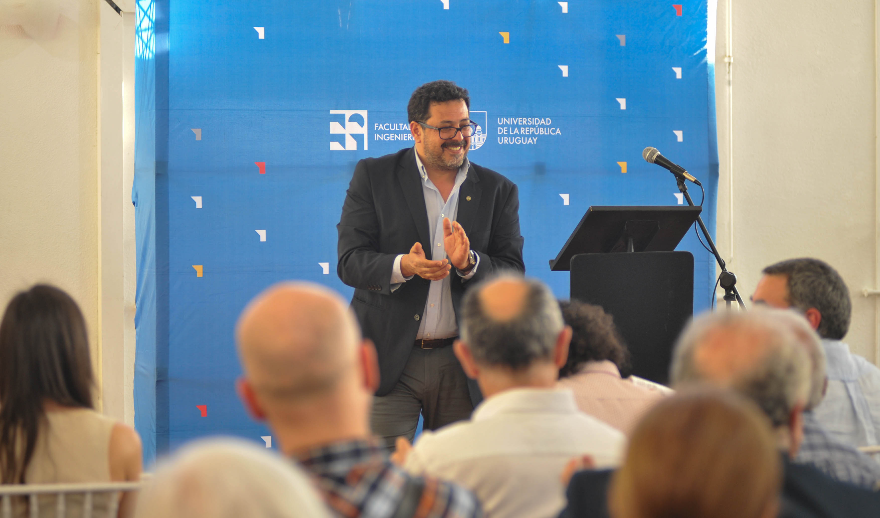 Secretario de Presidencia, de pie en un estrado, sonríe y aplaude frente al público. Detrás de él hay un panel azul con logo de la Udelar.