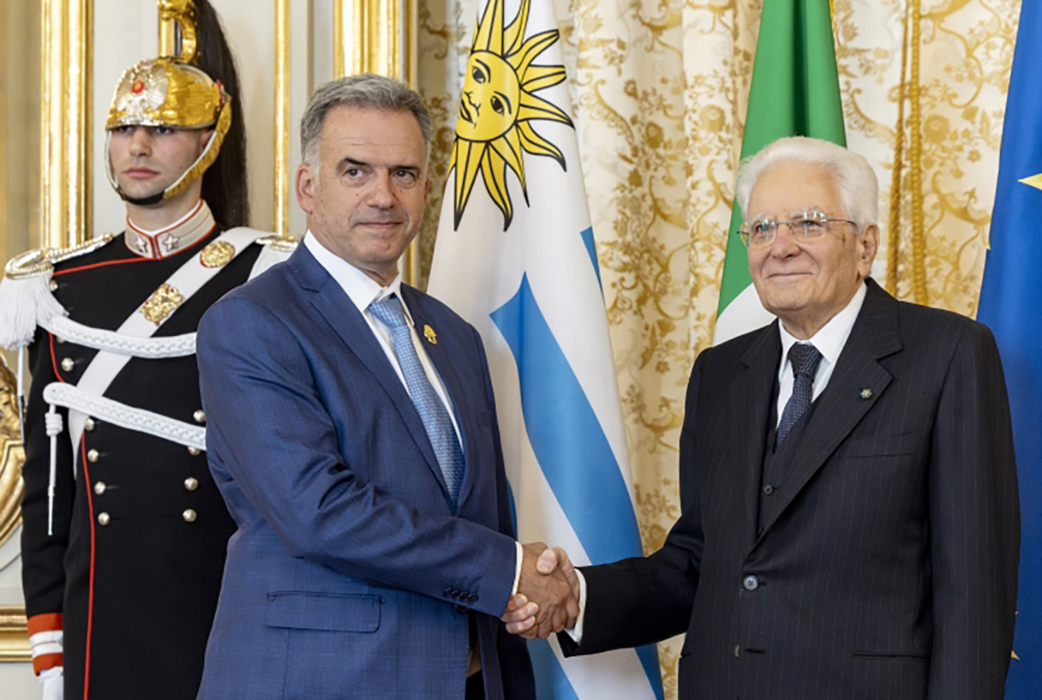 Los presidentes de Uruguay e Italia se estrechan la mano y sonríen a la cámara, con banderas de ambos países y un custodio de fondo