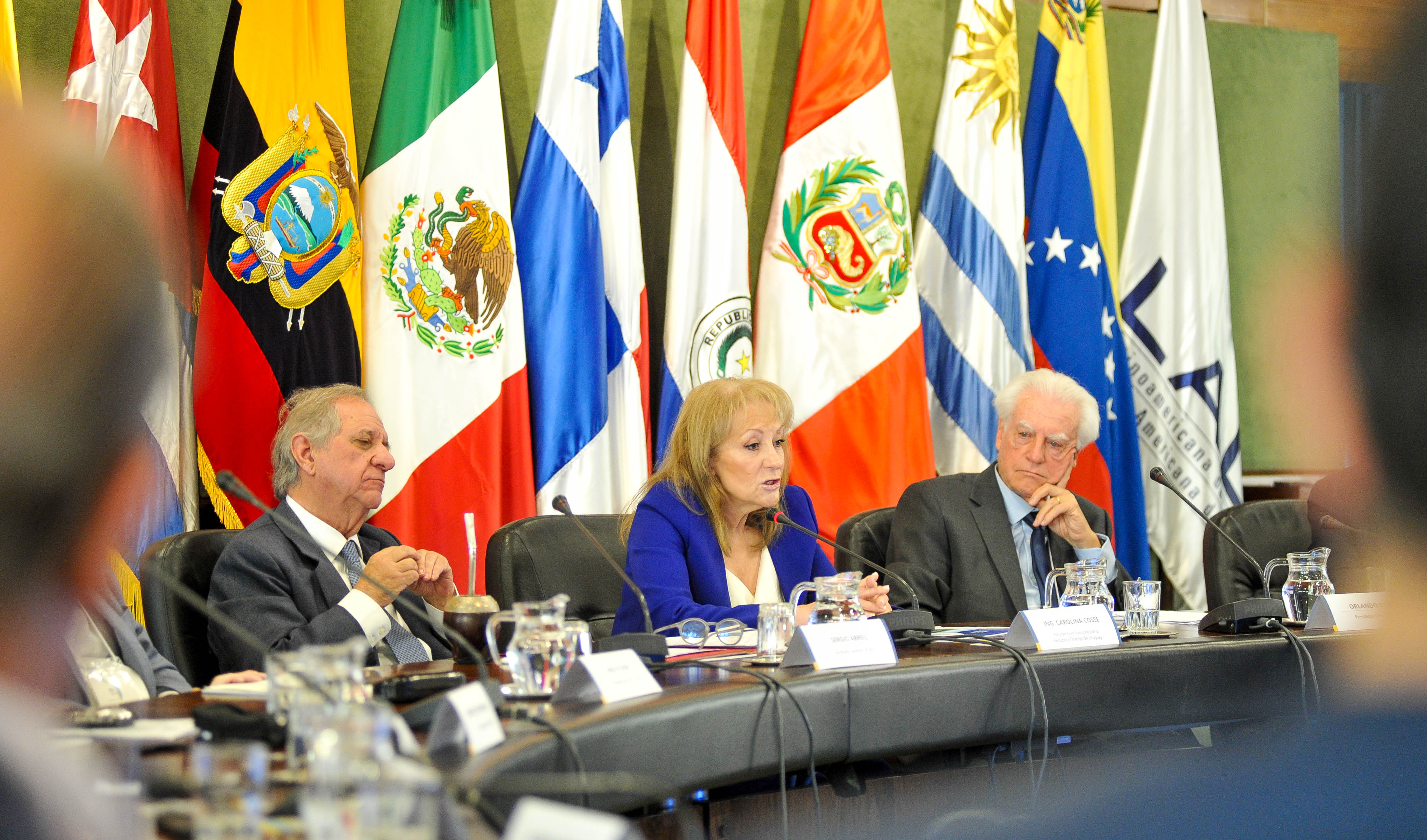 Cosse diserta sentada en mesa con micrófono junto a las autoridades de Aladi. Atrás se observan las banderas de varios países.