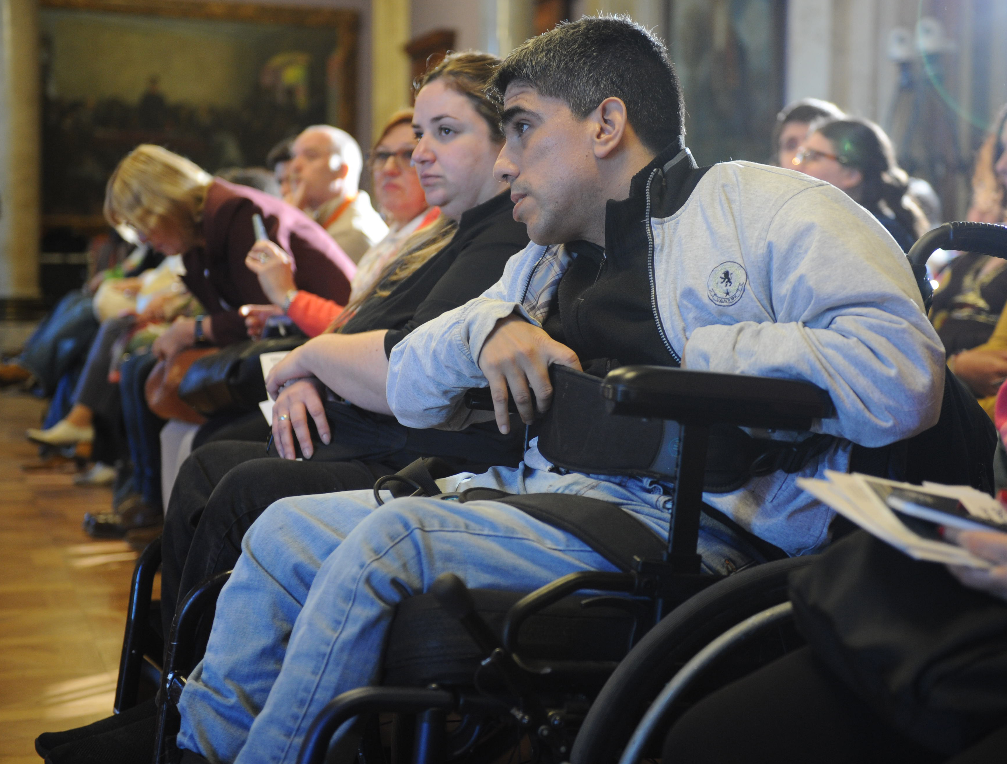 Dos usuarios de silla de ruedas sentados en primera fila junto al público miran hacia donde están disertando las autoridades.