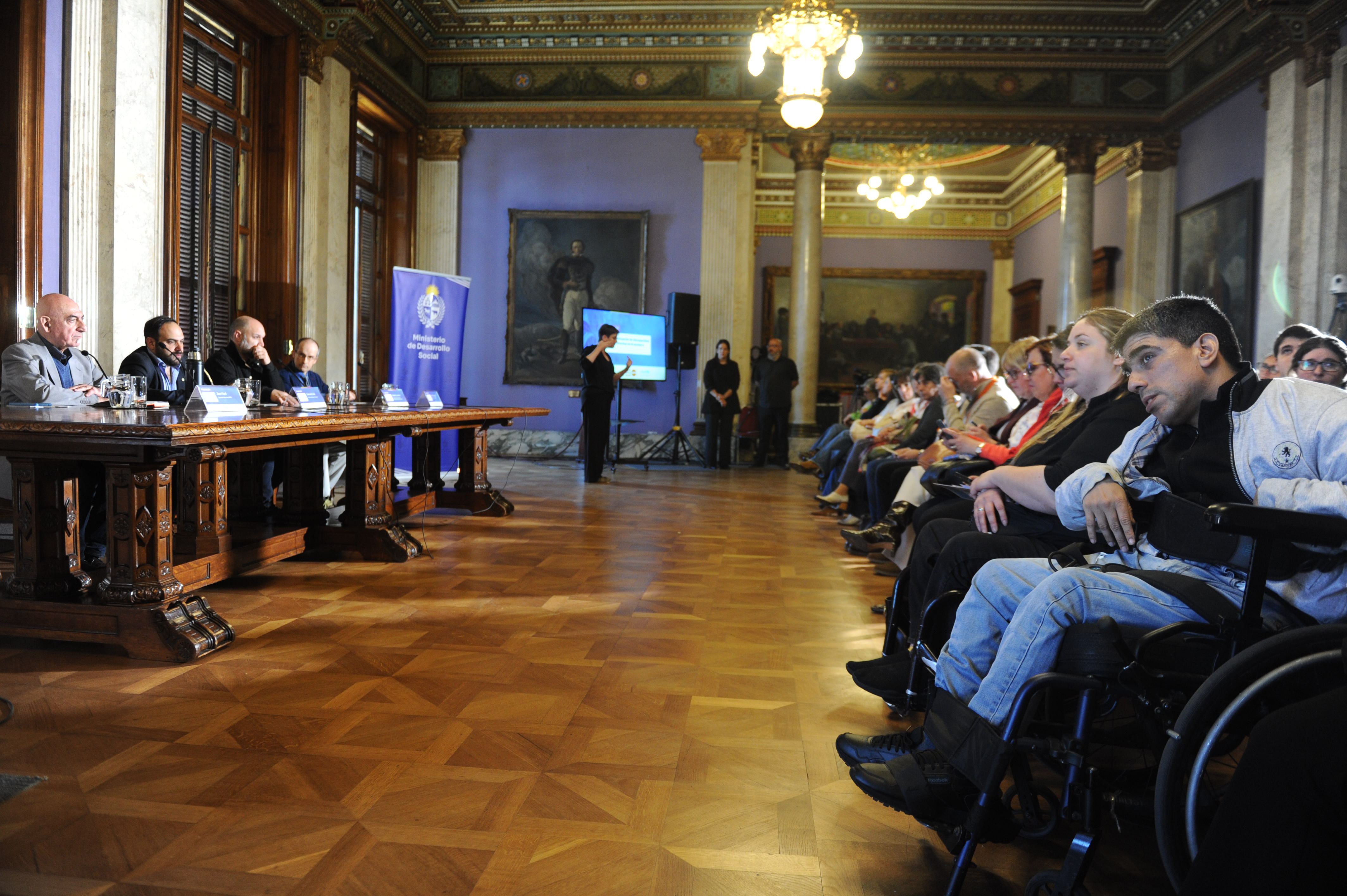 Plano general, de perfil, del público sentado en la platea y la mesa con autoridades disertando.