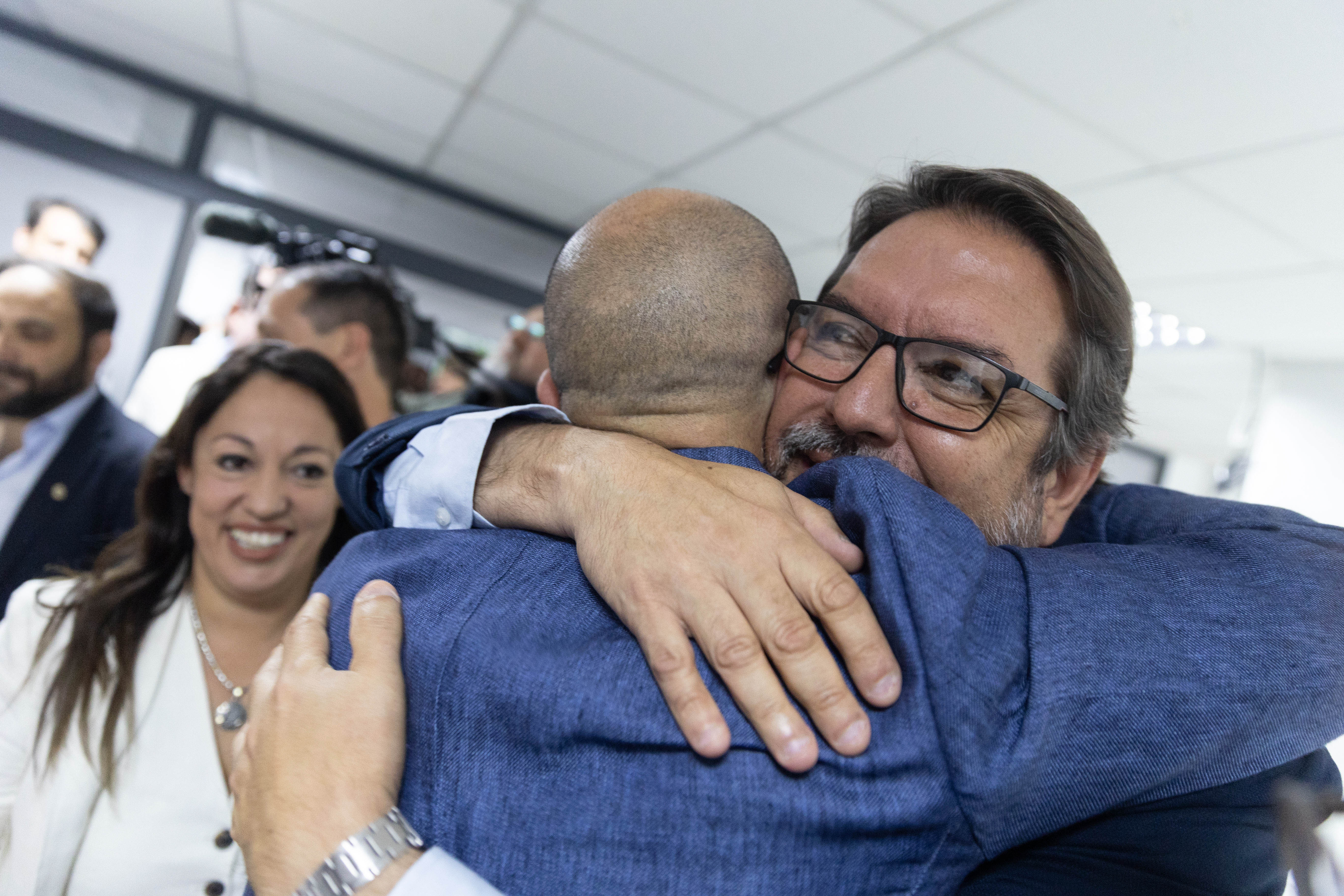 Primer plano de dos hombres abrazándose mientras la ministra de Industria los observa.