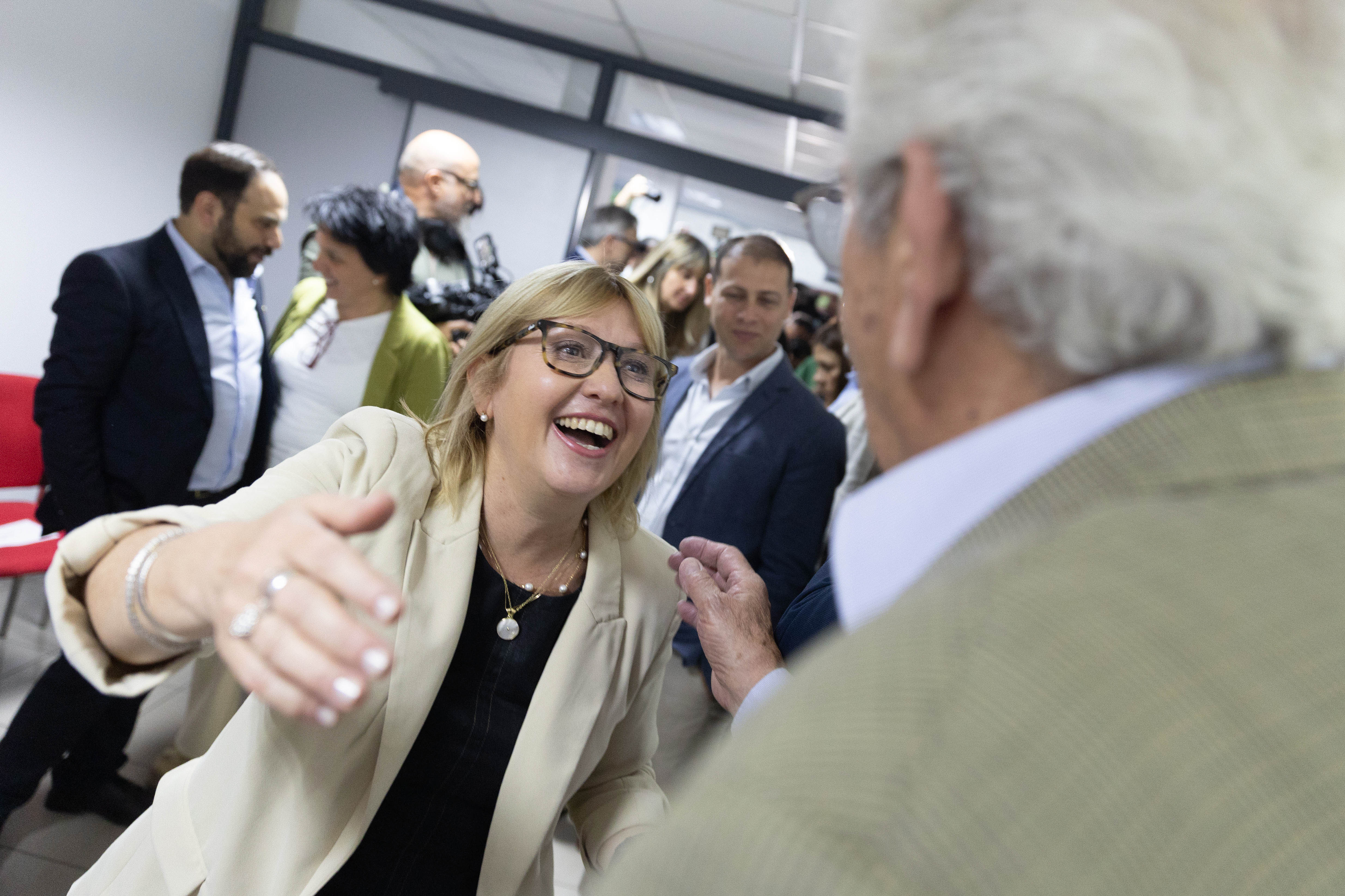 Primer plano de la ministra de Salud Pública, sonriendo y extendiendo los brazos para saludar a un hombre. Atrás hay otras personas conversando.