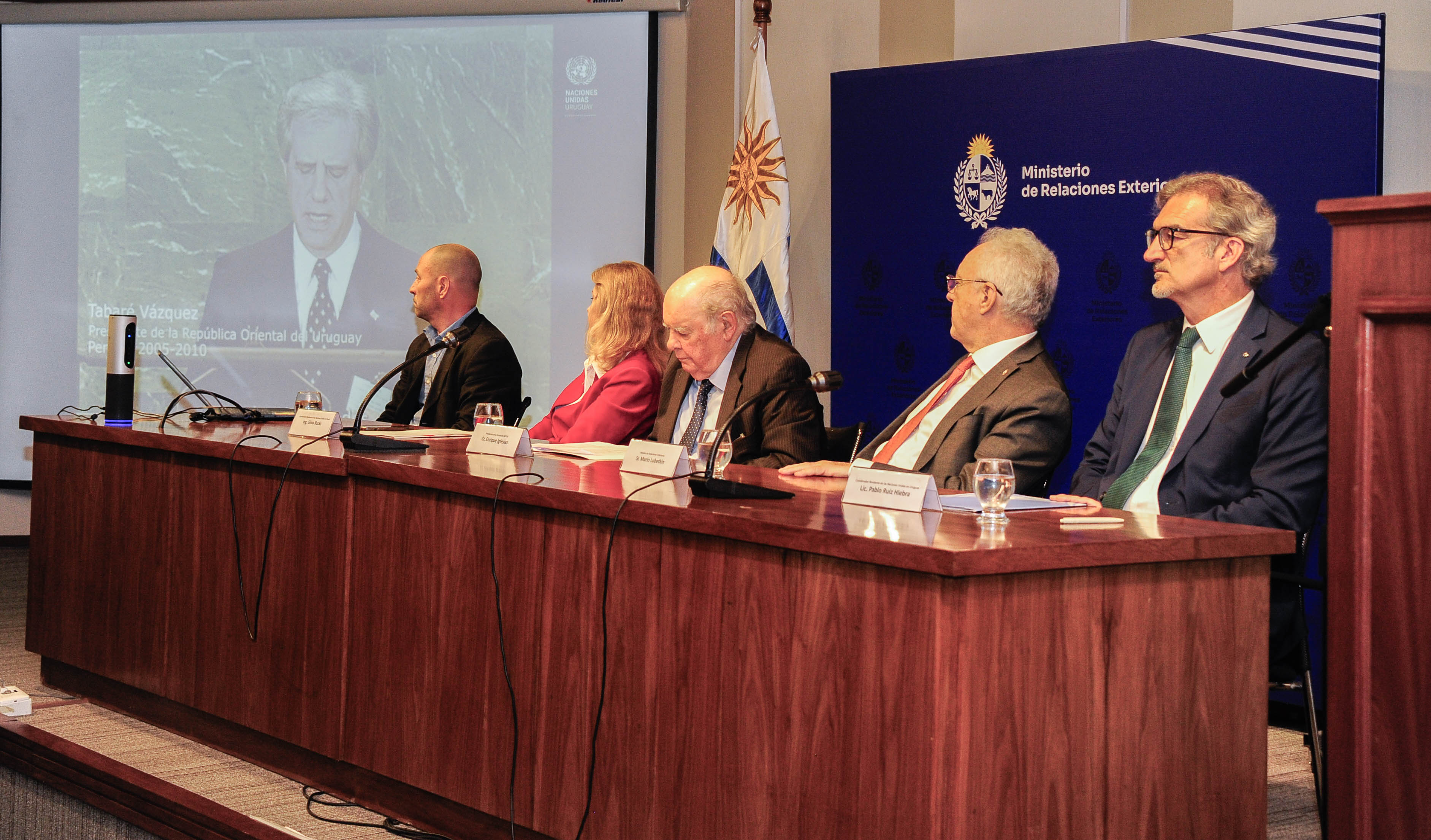 Cinco autoridades sentadas en mesa con micrófono miran hacia una gran pantalla donde se emite un video del expresidente Tabaré Vázquez.
