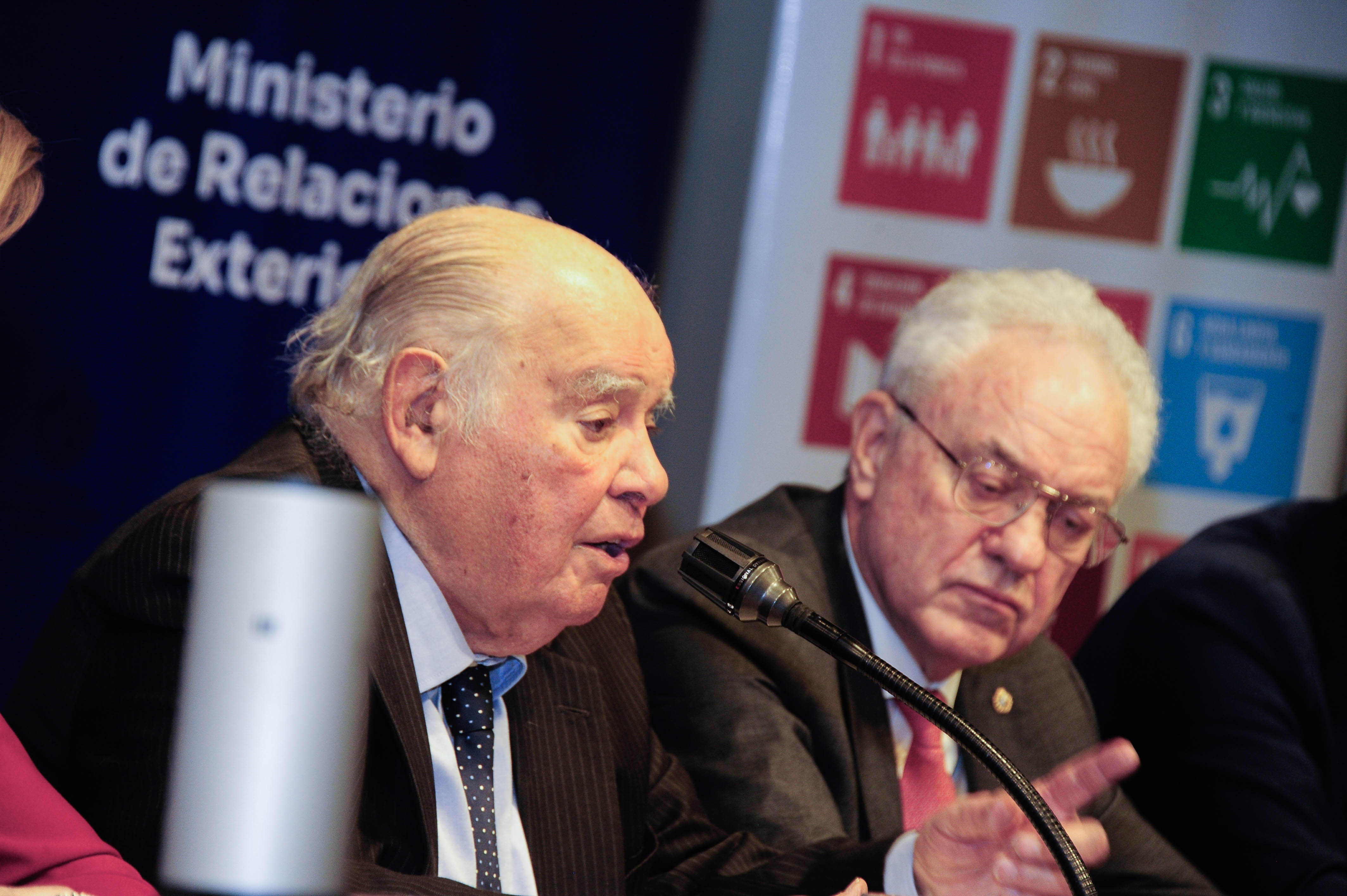Plano lateral del presidente de la Fundación Astur disertando sentado en mesa con micrófono mientras el canciller lo observa.