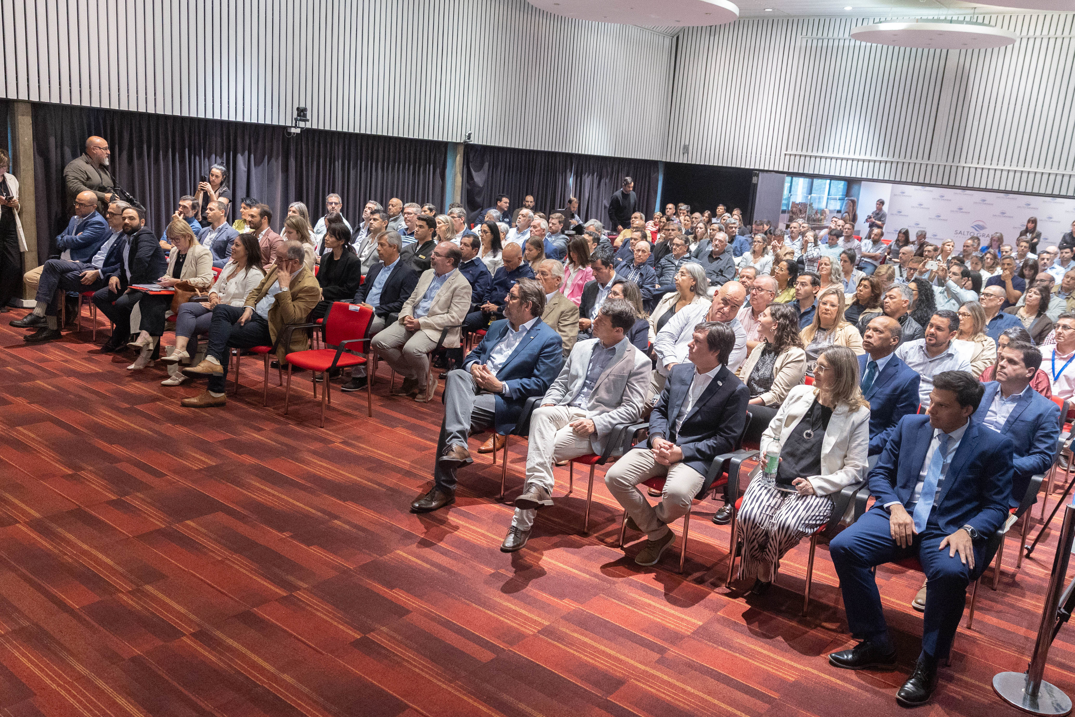 Plano general del salón de actos con el público sentado en la platea escuchando una disertación.