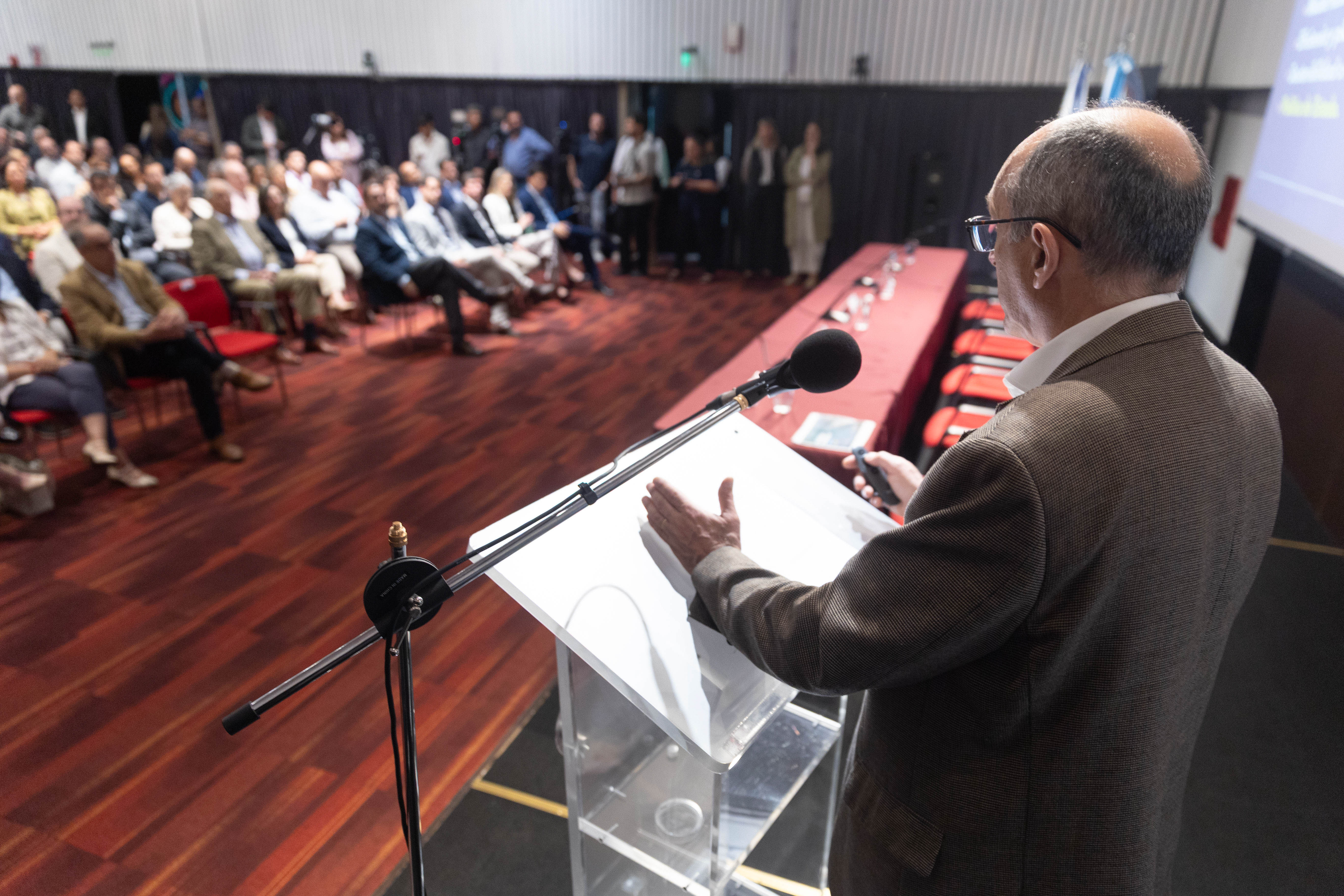 El presidente de la CTM por Uruguay, de espaldas, diserta de pie en atril con micrófono ante el público sentado en la platea del salón de actos.