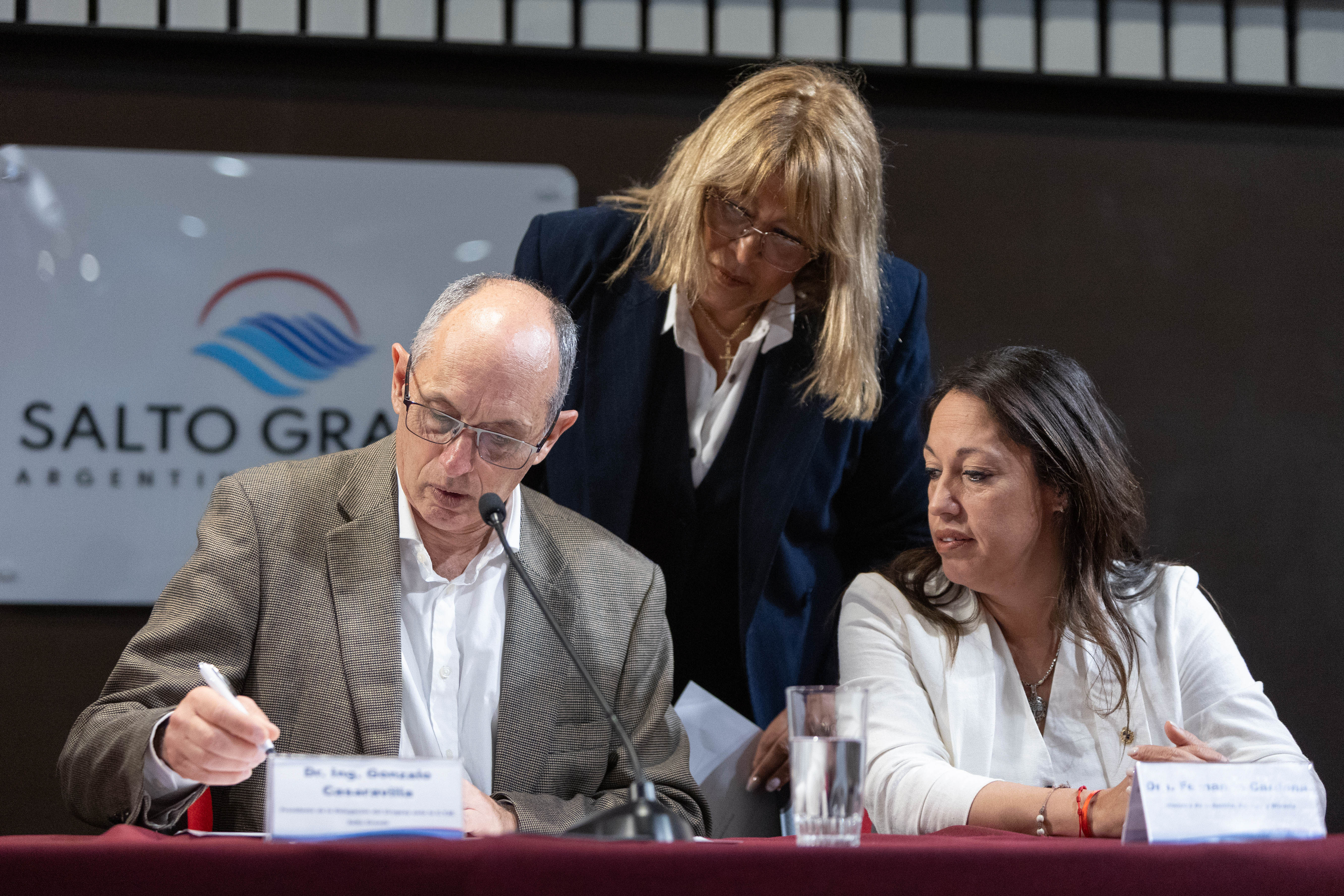 Primer plano del presidente de la CTM, sentado en mesa, firmando unos documentos. La ministra de Industria y una escribana lo observan.