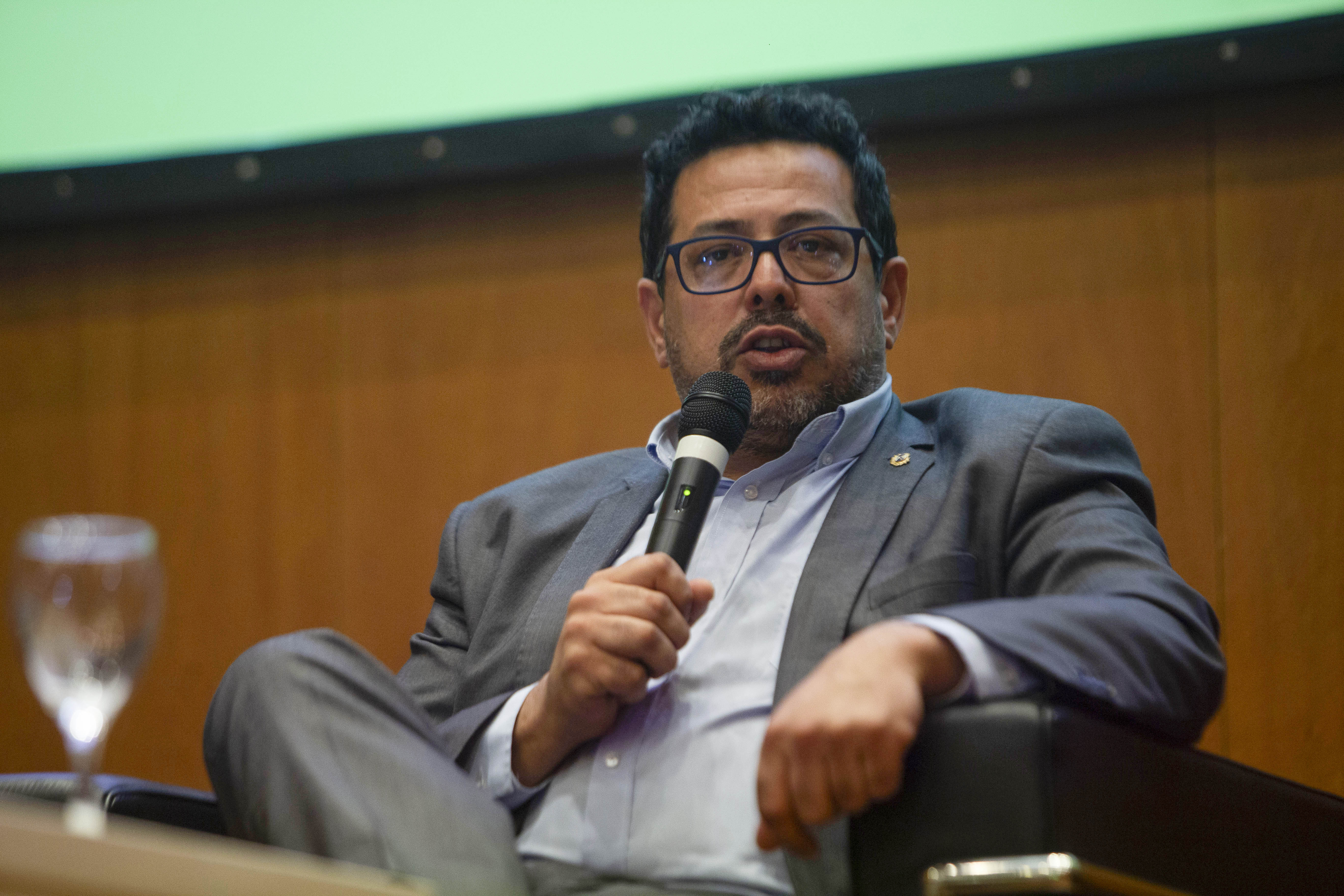 Secretario de Presidencia diserta, sentado, con micrófono en mano en el auditorio de Presidencia.