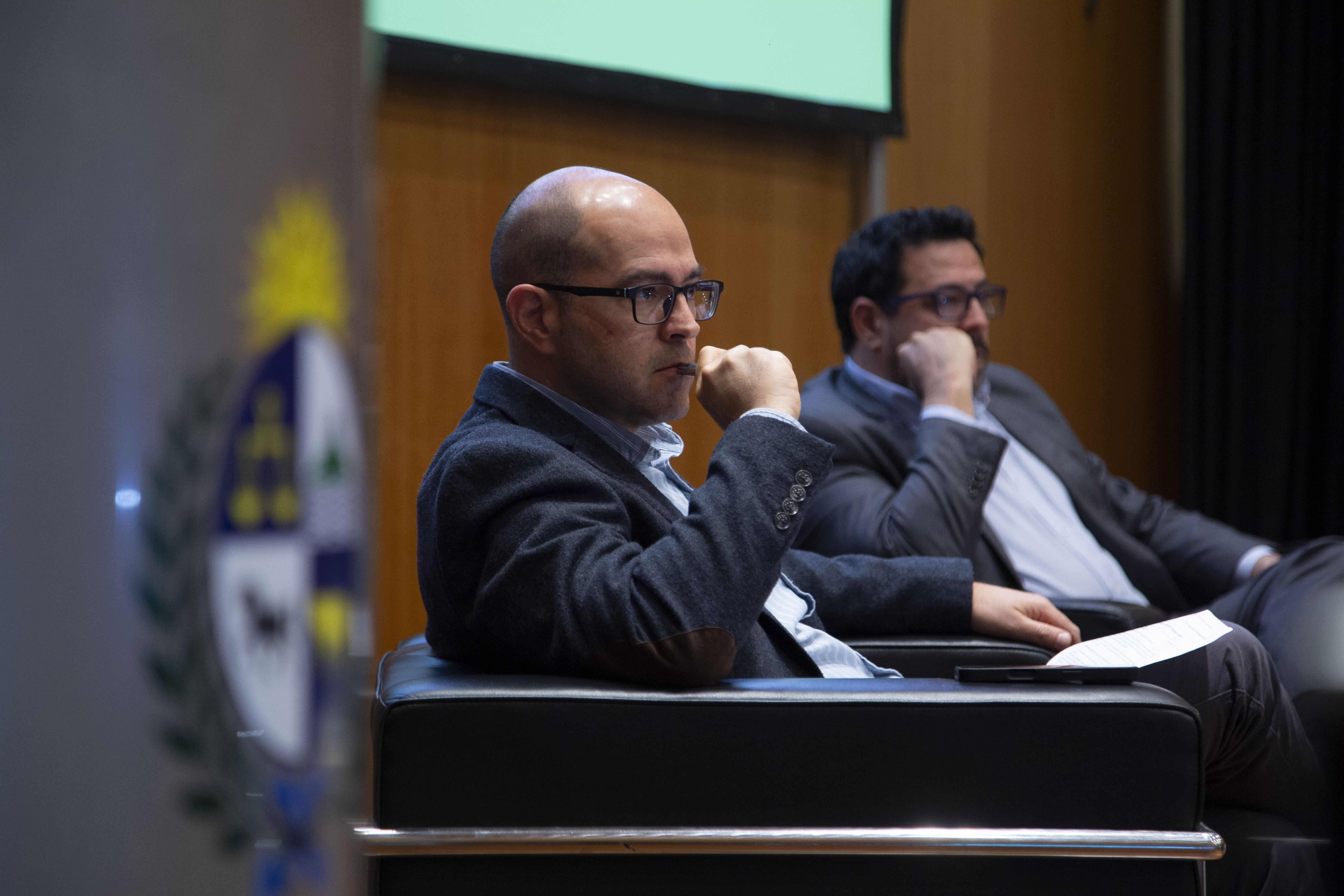 Secretario de Presidencia y presidente de la ASSE escuchan a un disertante, sentados en el escenario.