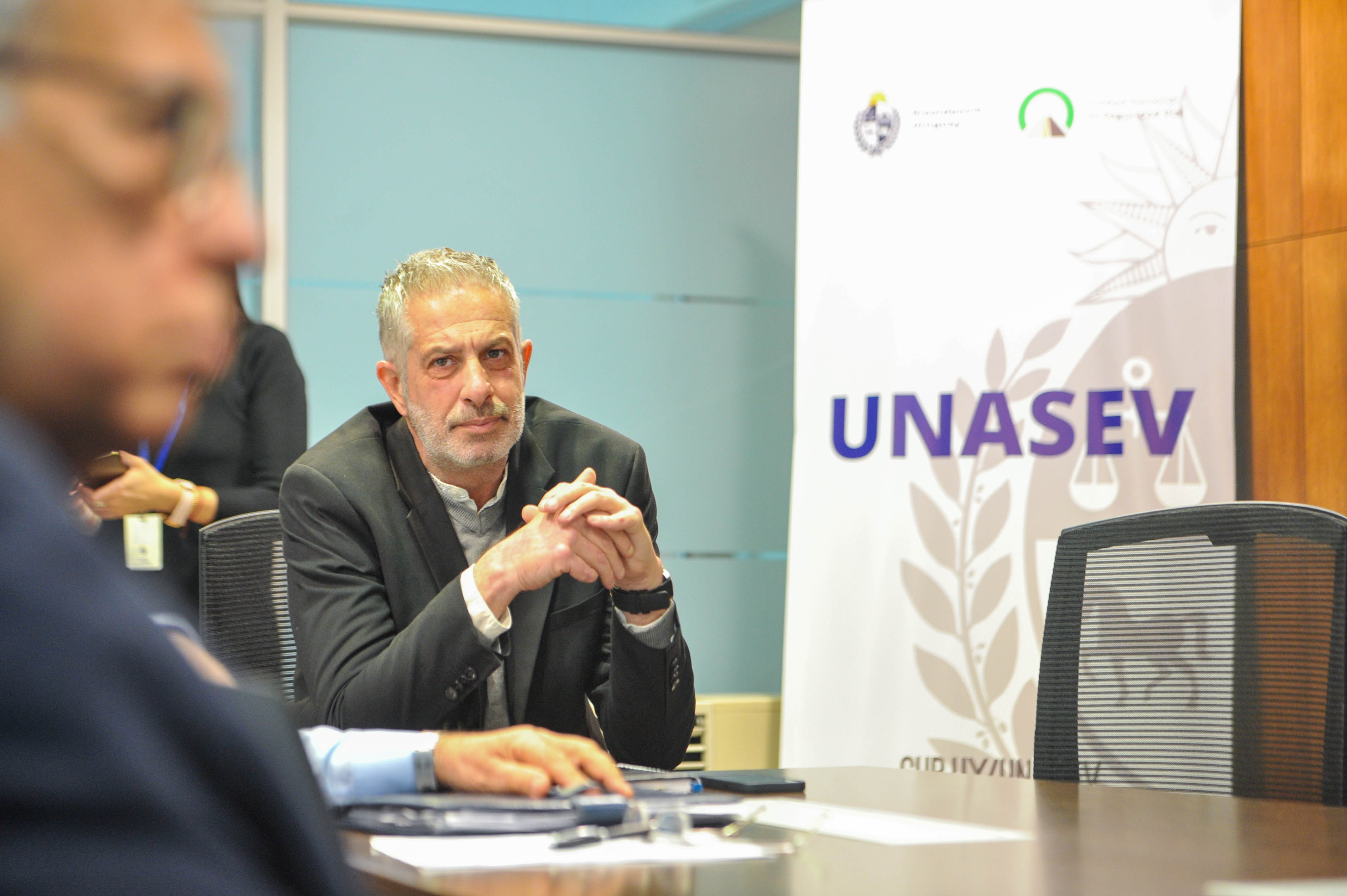 Un representante municipal sentado en la mesa mira hacia un lado. Atrás se observa cartel blanco con logo de Unasev.