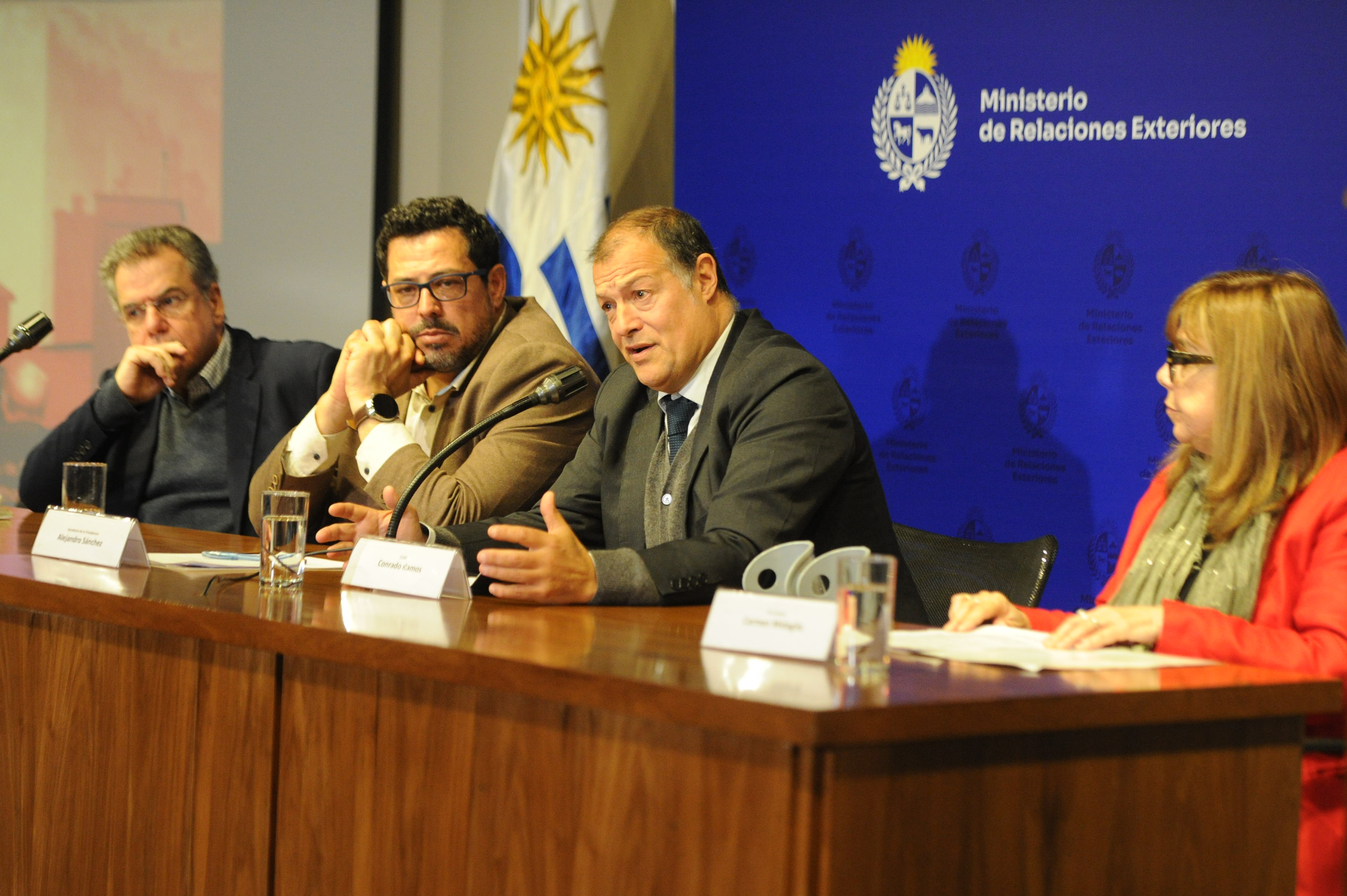 El secretario general del CLAD diserta sentado en mesa con micrófono junto al secretario de Presidencia y otros dos oradores.
