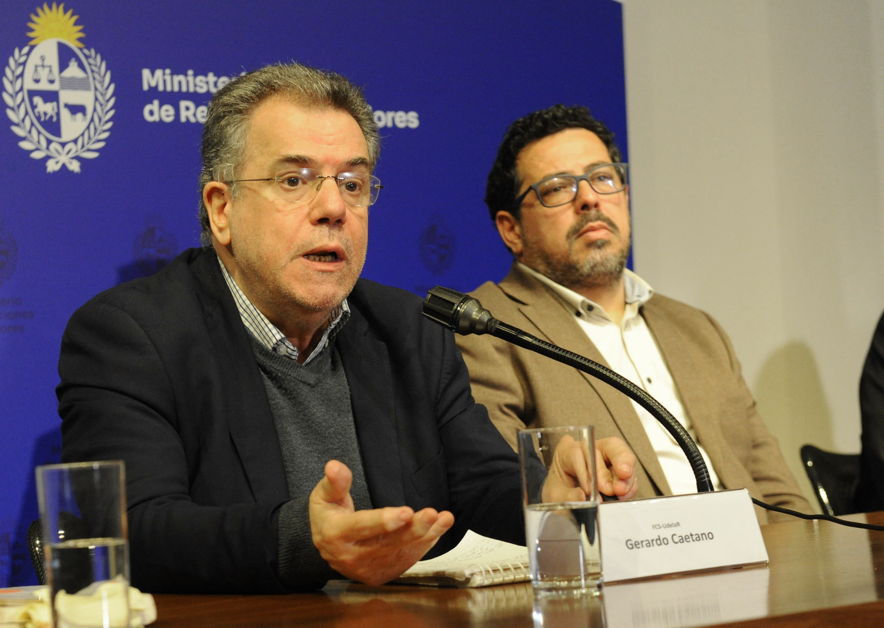 El historiador Gerardo Caetano diserta sentado en mesa con micrófono junto al secretario de Presidencia. Atrás hay un cartel azul con logo del MRREE.