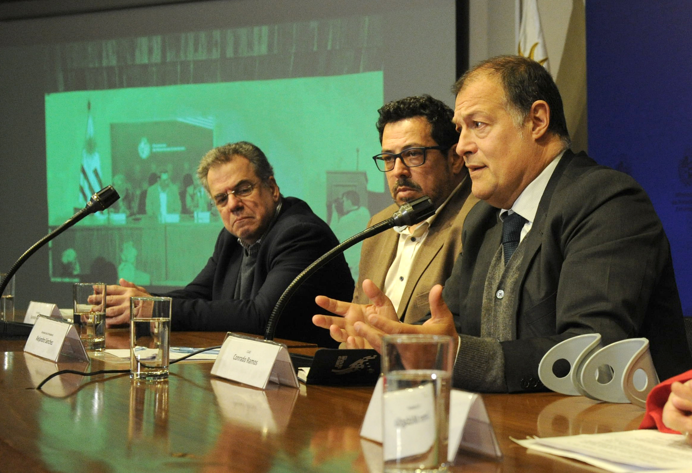 El secretario general del CLAD diserta sentado en mesa con micrófono junto al secretario de Presidencia y el historiador Caetano.