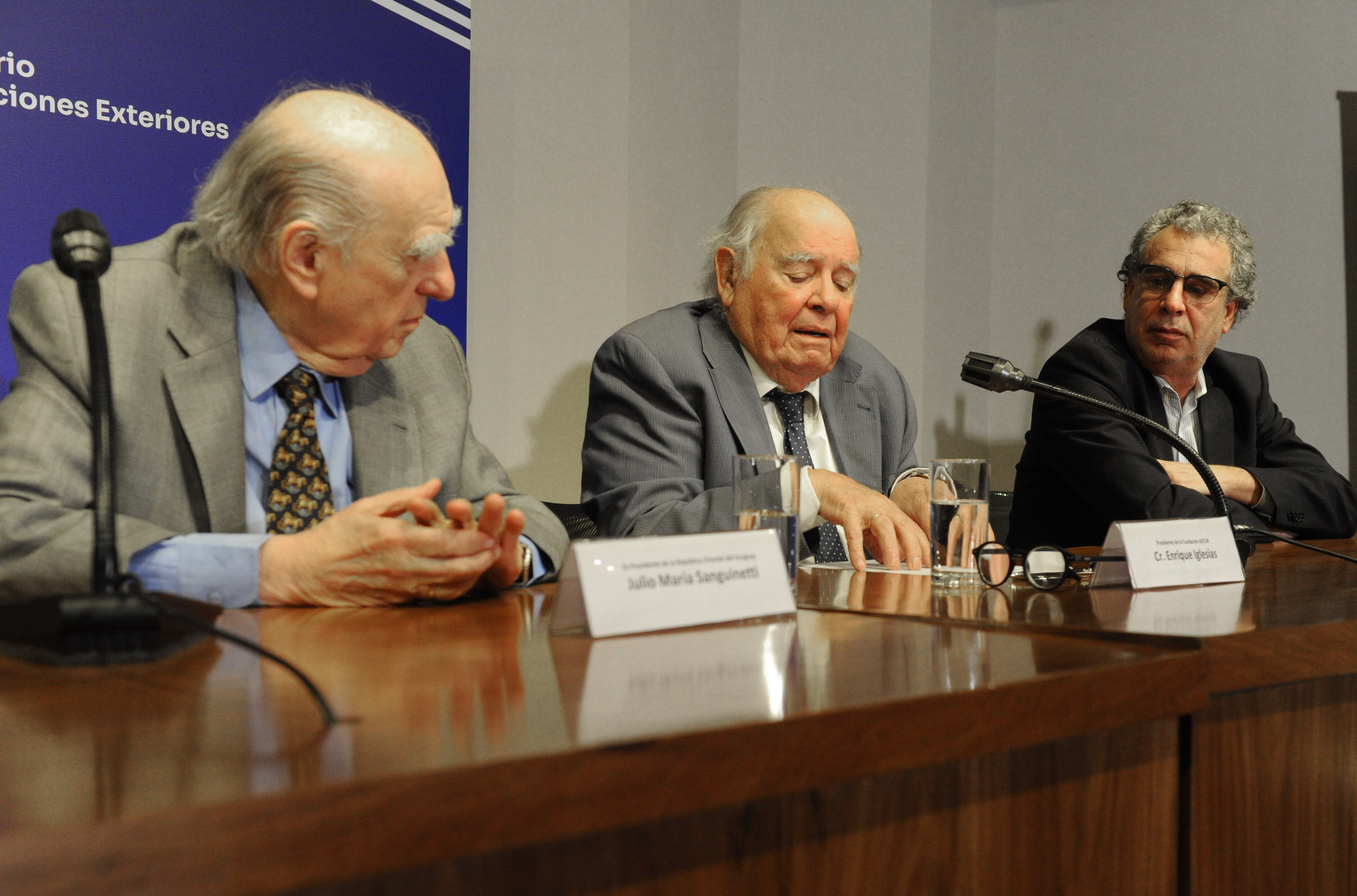 Exsecretario ejecutivo de la Cepal diserta sentado en mesa con micrófono. A sus costados dos oradores lo observan.