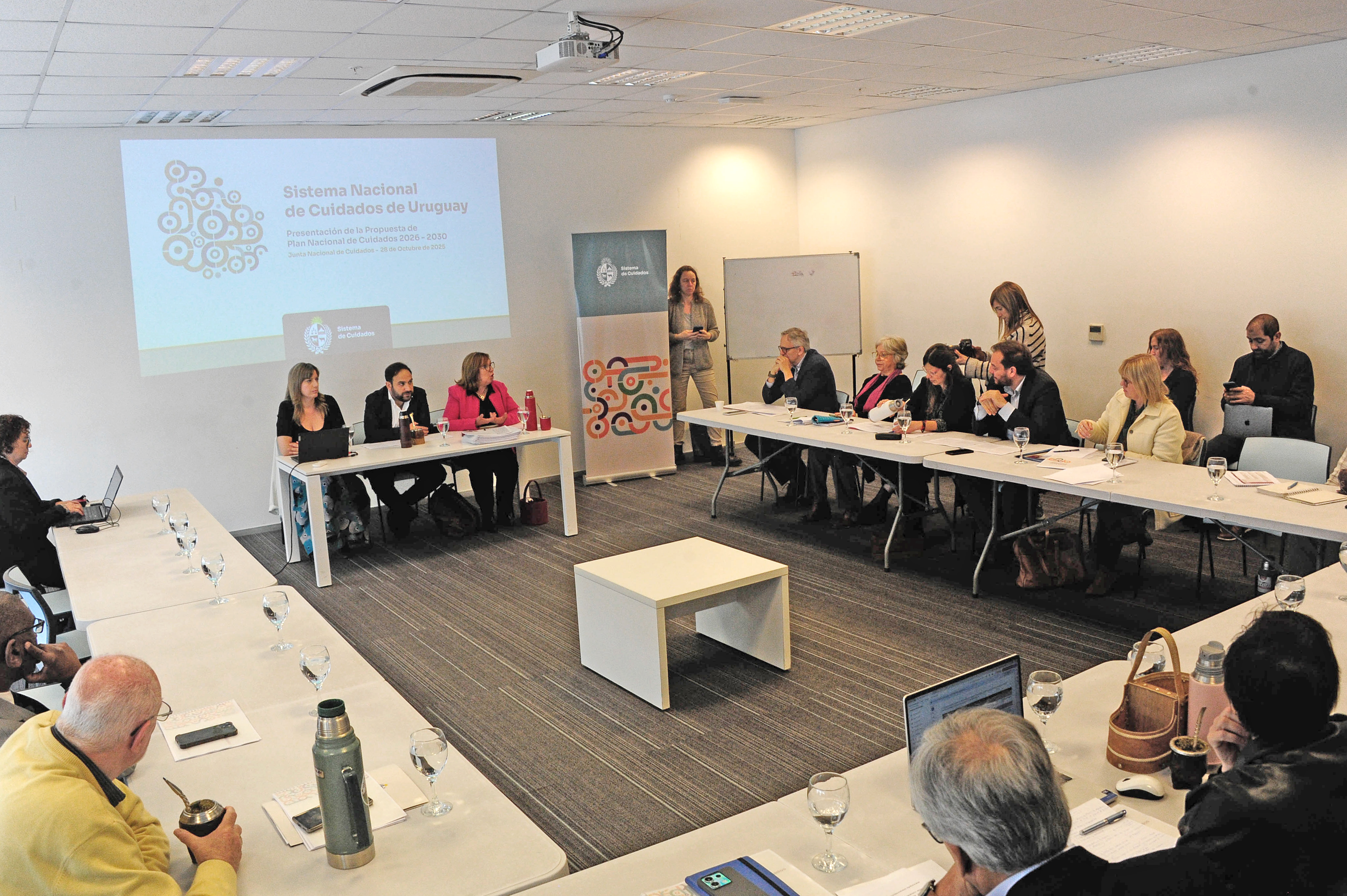 Plano general de la sala de reuniones con las autoridades sentadas en mesas en forma de U dialogando.