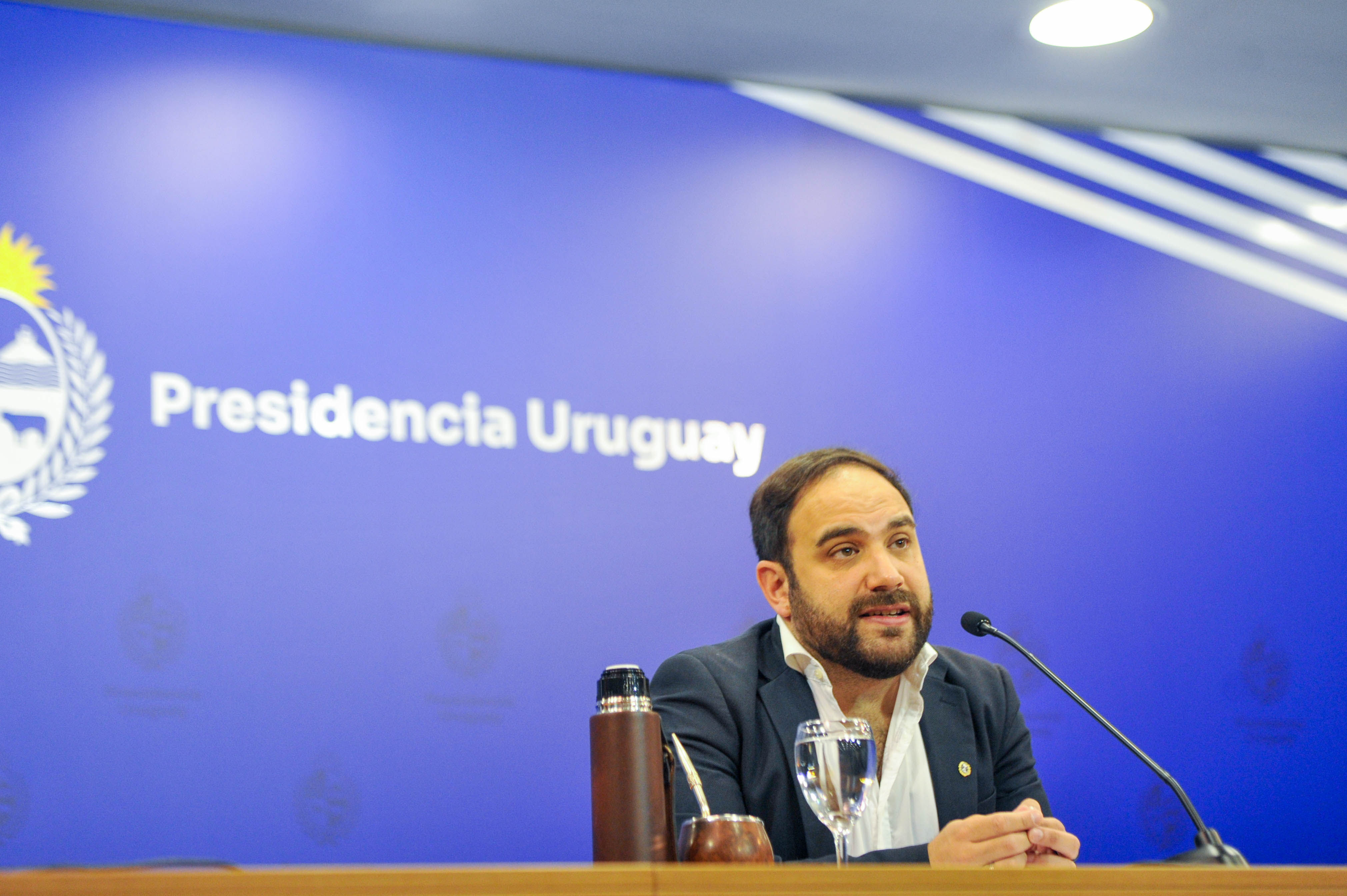 Ministro de Desarrollo Social diserta sentado en mesa con micrófono en sala de prensa de Torre Ejecutiva. Atrás hay un cartel azul con logo de Preside