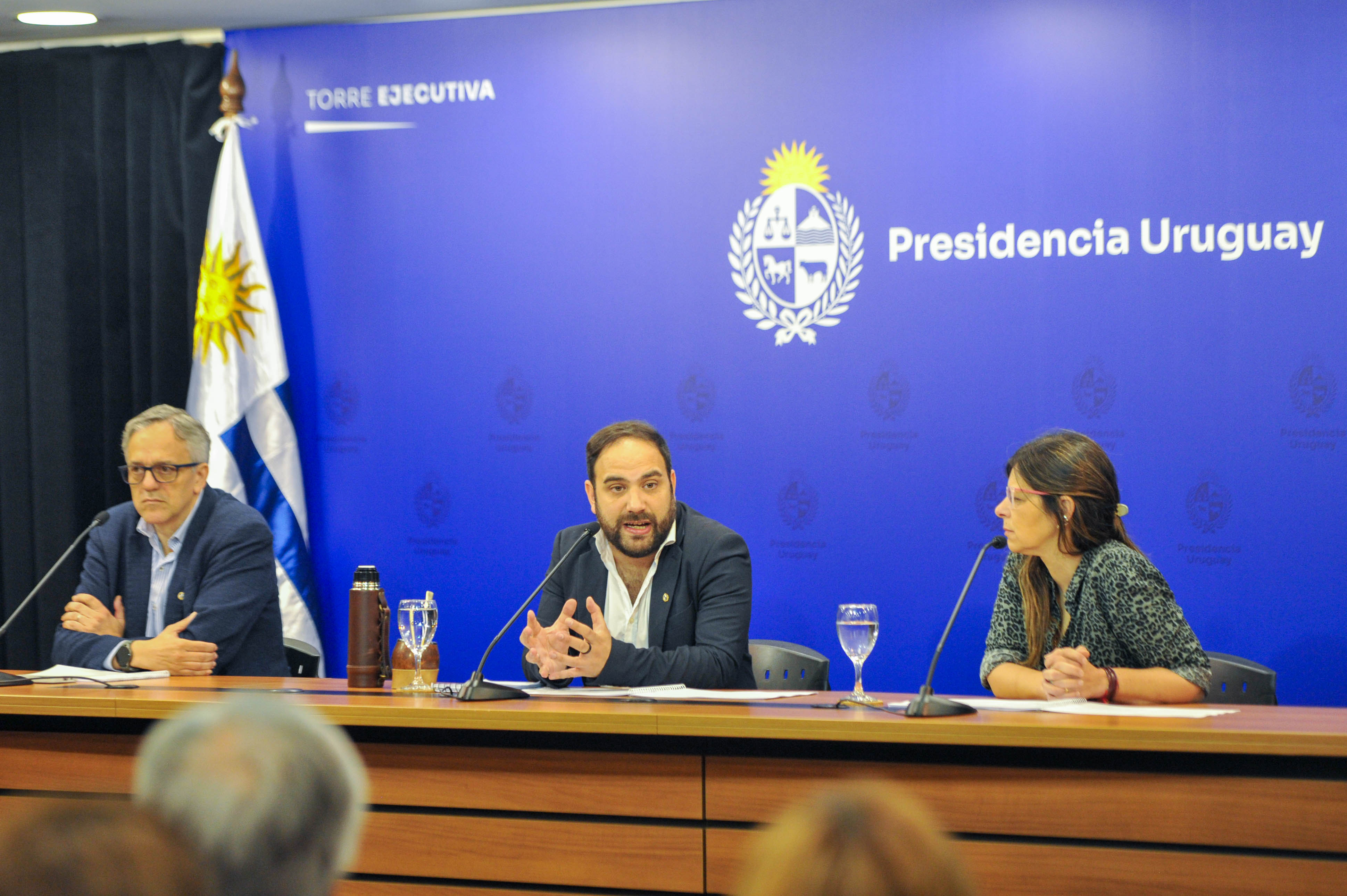 Ministro de Desarrollo Social diserta sentado en mesa con micrófono junto a la presidenta del INAU y el director de OPP.