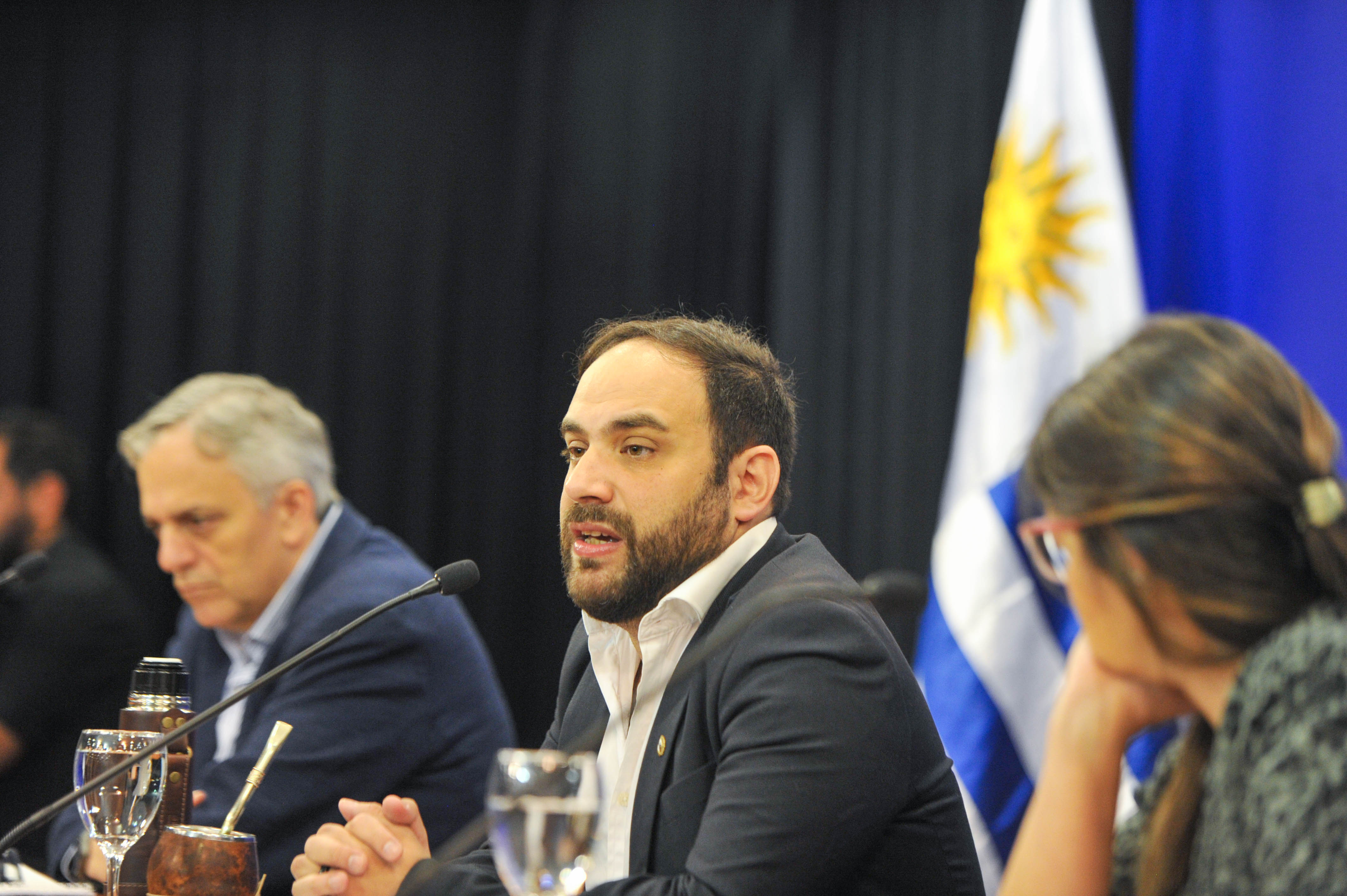 Ministro de Desarrollo Social diserta sentado en mesa con micrófono, mientras la presidenta del INAU y el director de OPP lo observan.