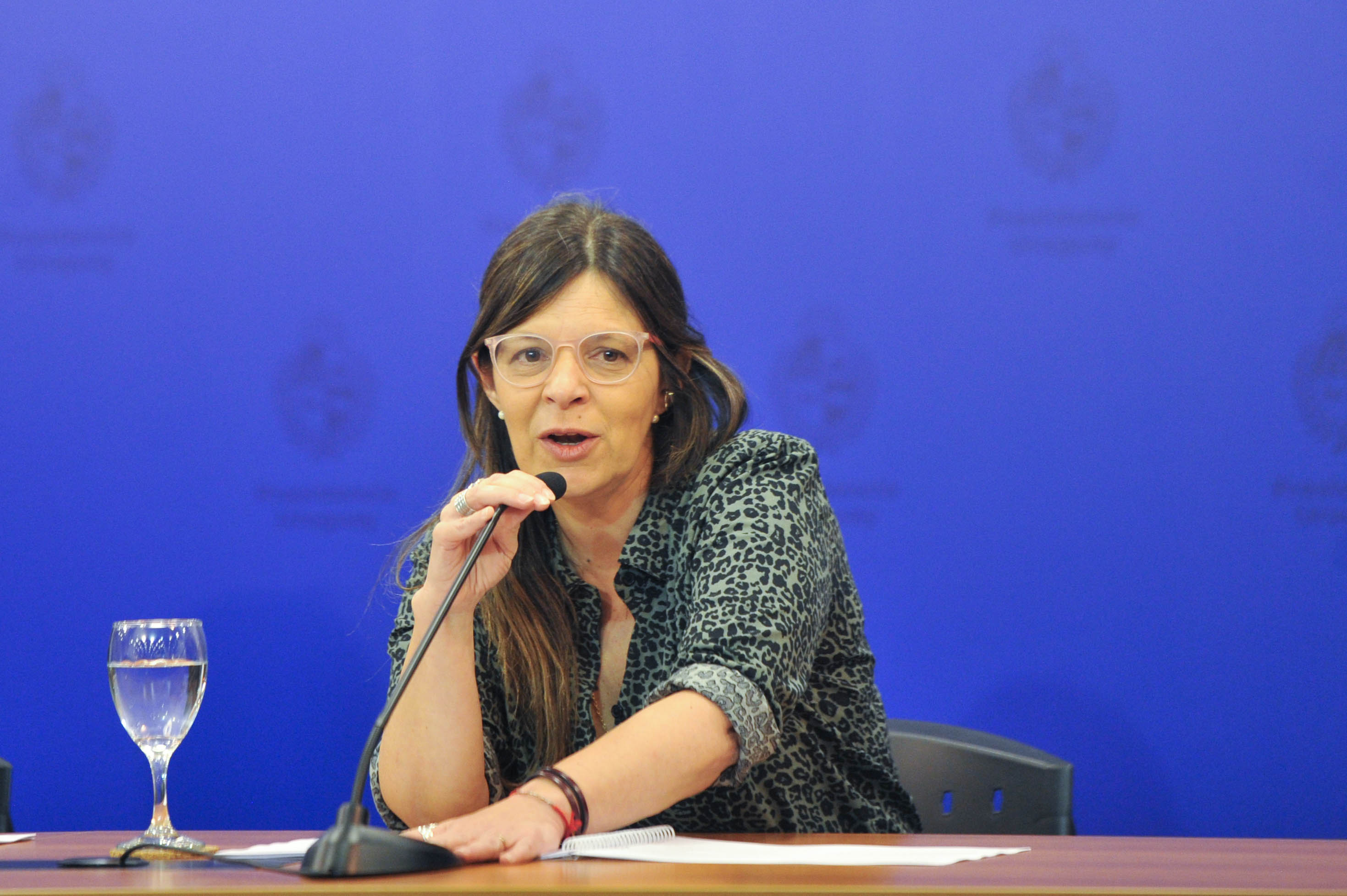 Presidenta de INAU diserta sentada en mesa con micrófono en sala de prensa de Torre Ejecutiva. Atrás hay un cartel azul con logo de Presidencia.