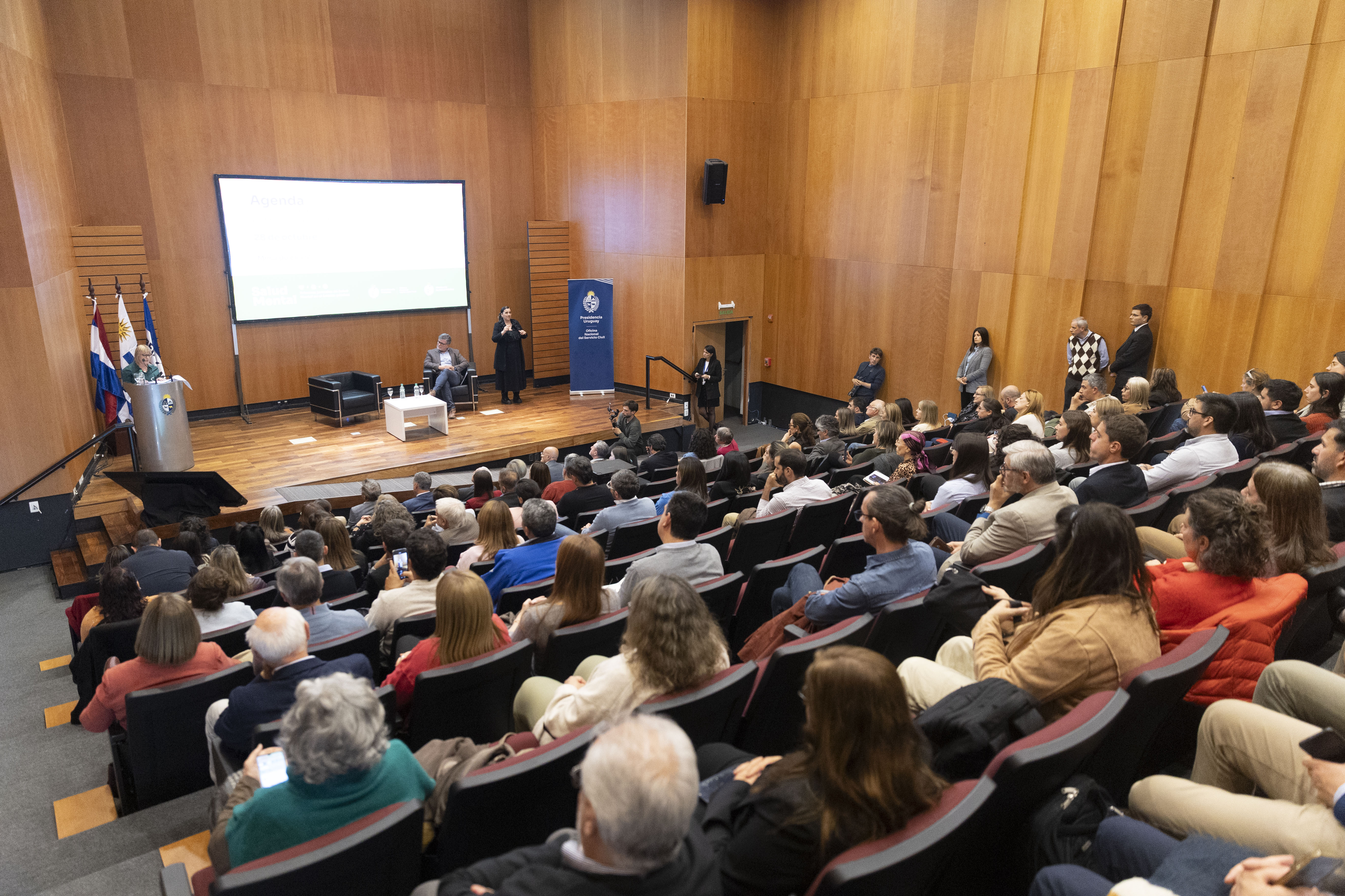 Plano general del auditorio, con los asistentes observando a la ministra de Salud Pública, quien diserta de pie en un atril.
