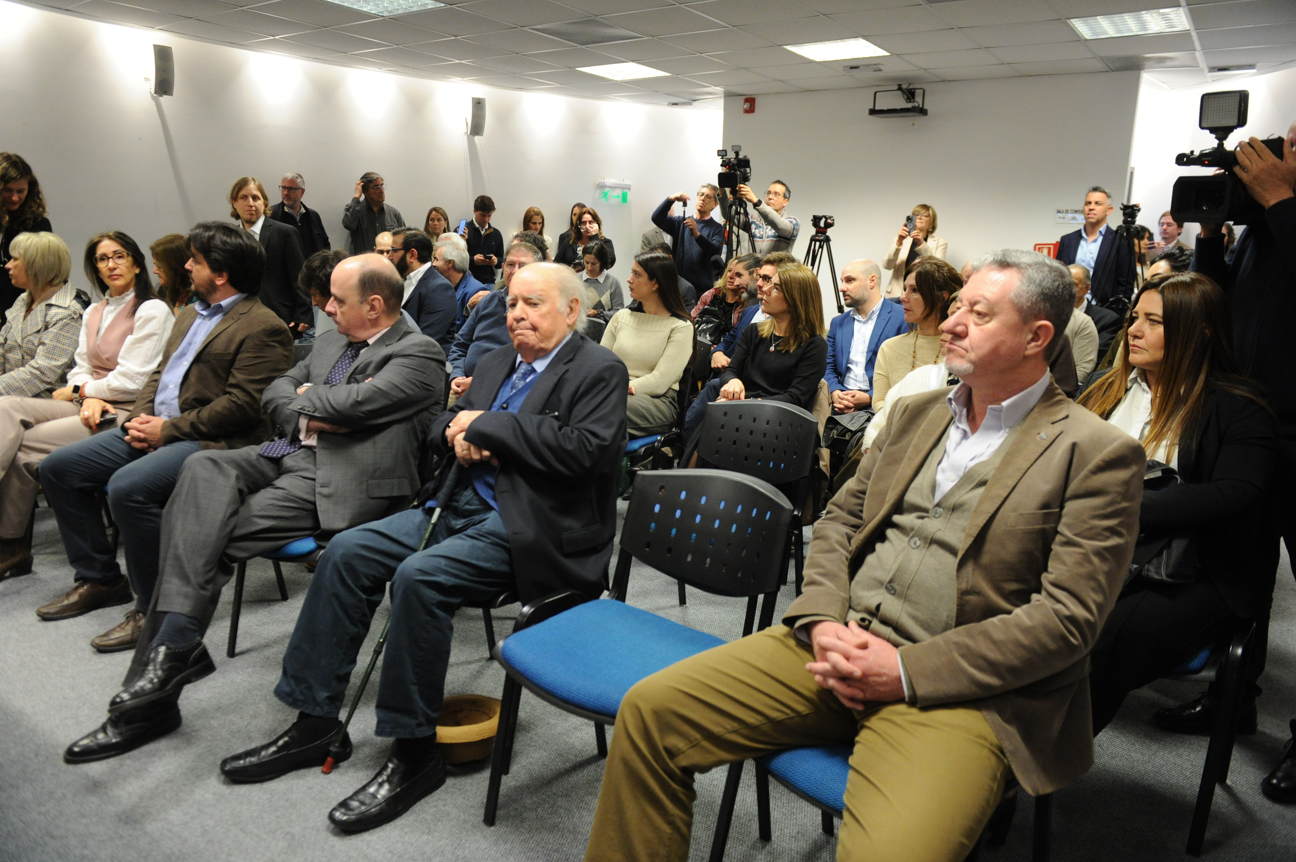 Asistentes, sentados en la platea, escuchan con atención la conferencia. Se observan cámaras al fondo.